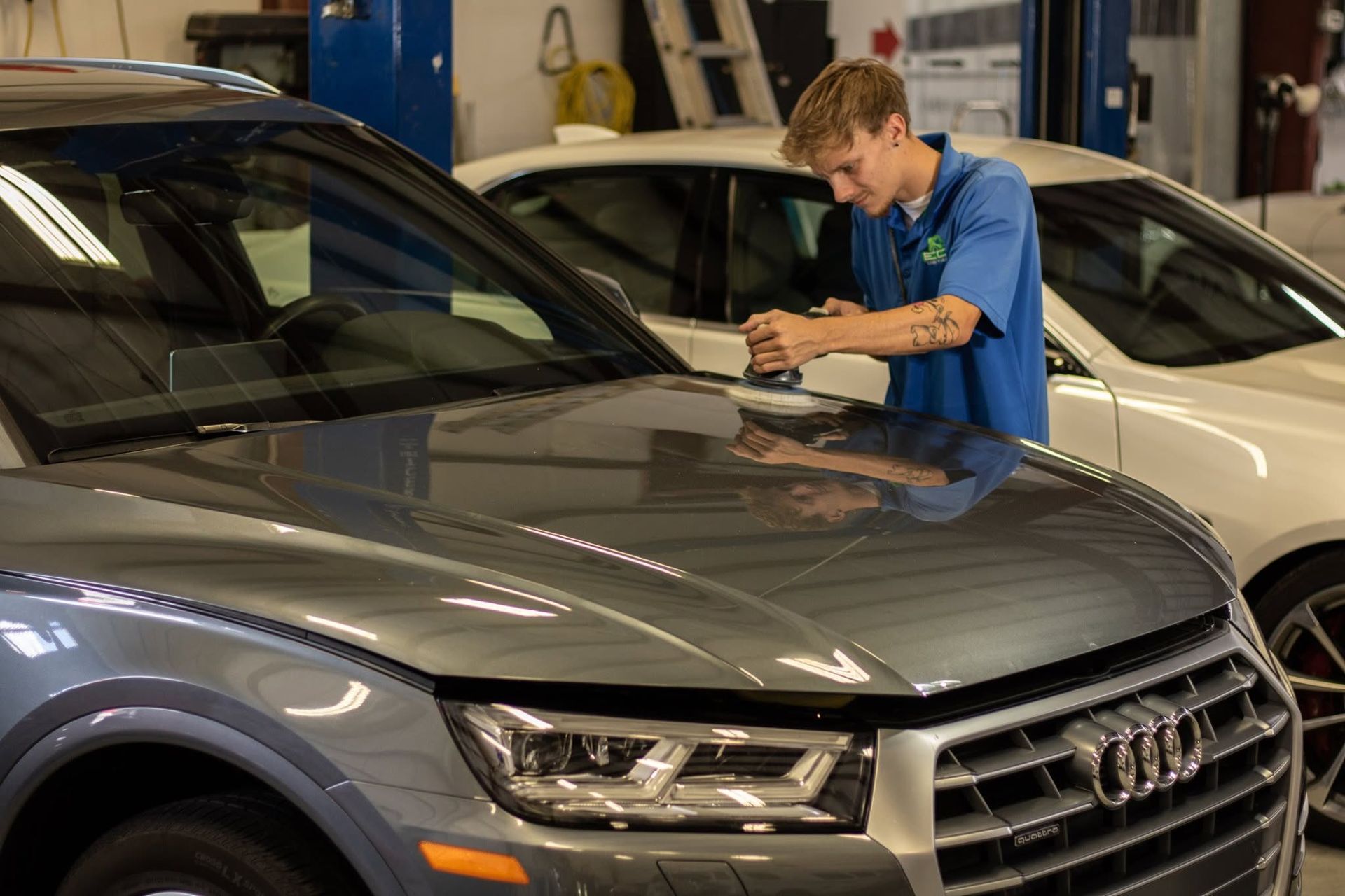 Mechanic inspecting a gray Audi in an auto shop, with a white car in the background