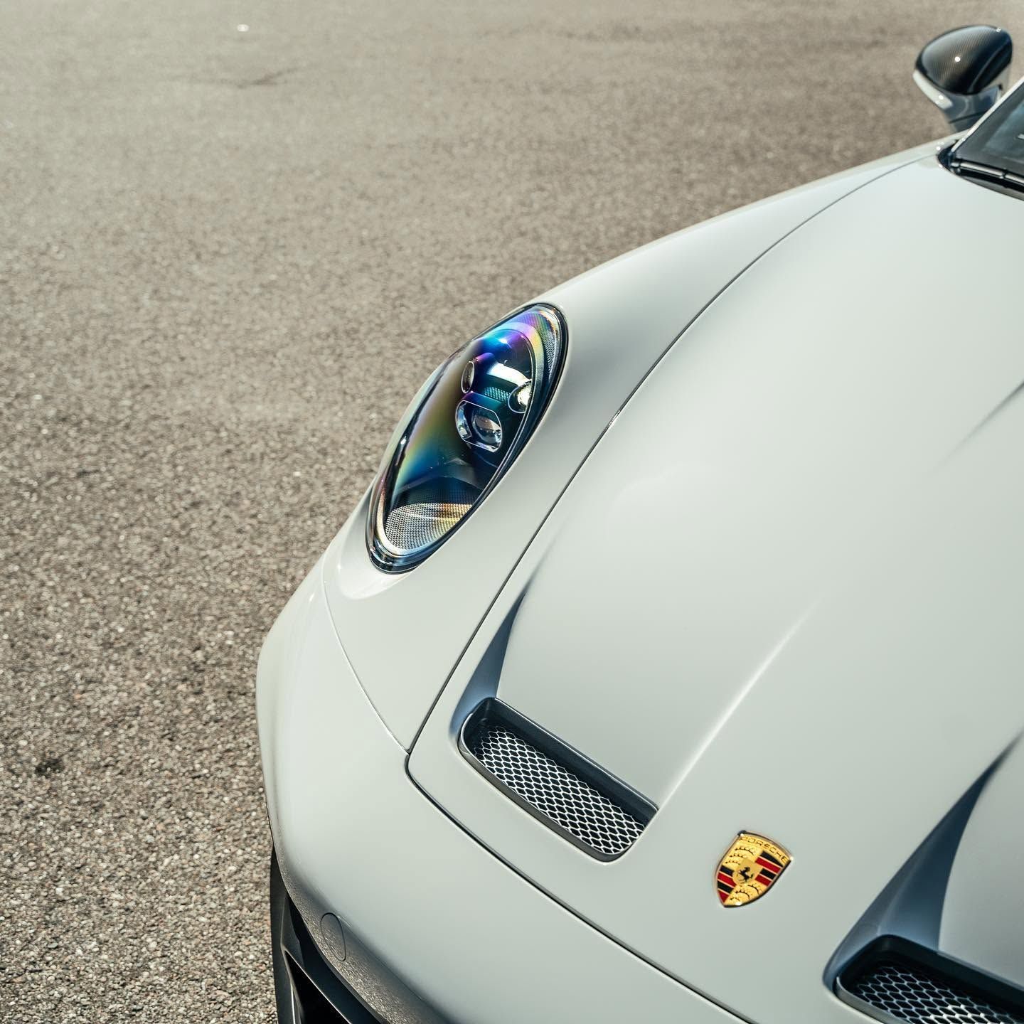 Close-up of a white Porsche sports car front end on asphalt, showing the headlight and hood badge