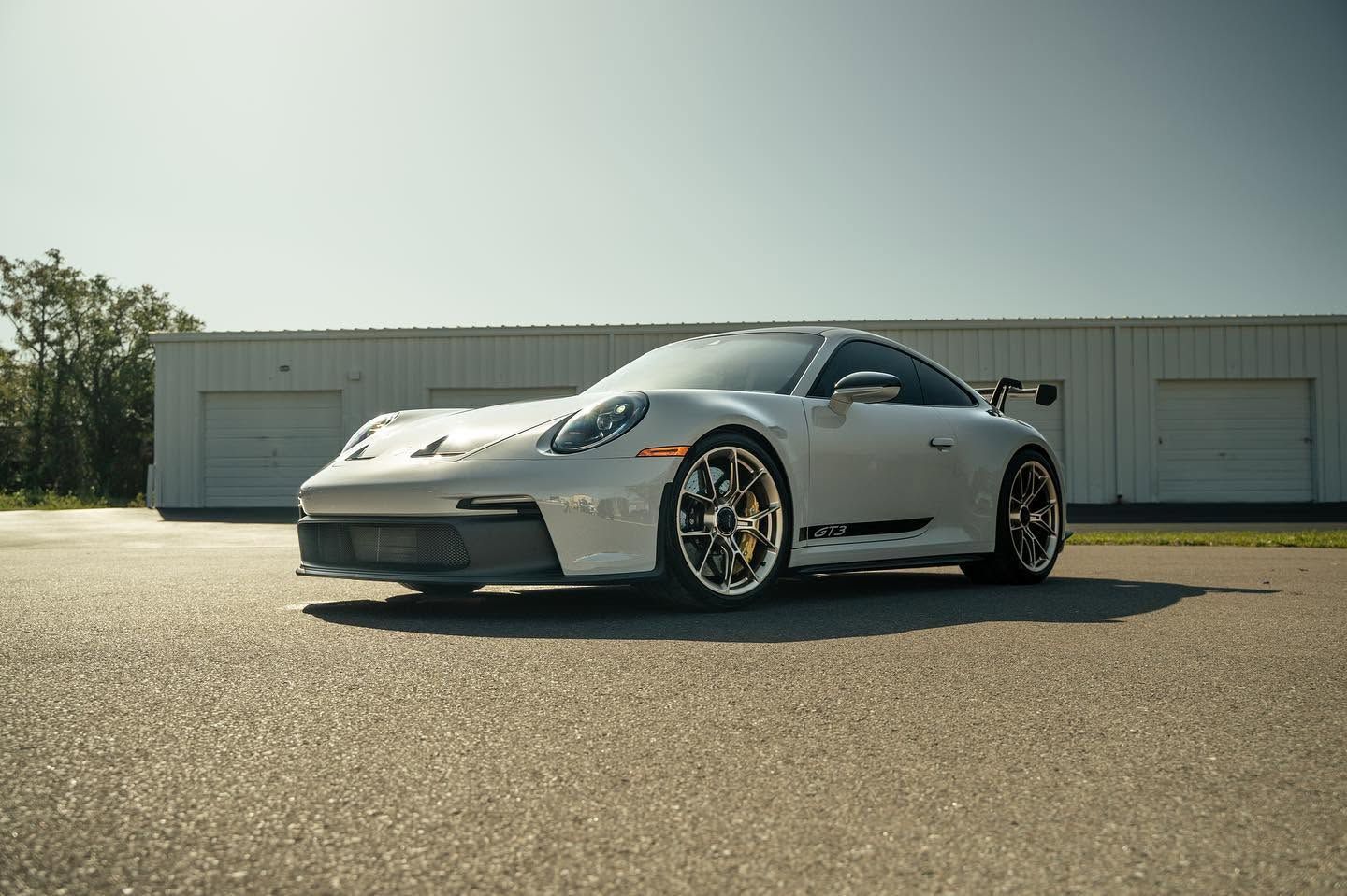 Silver sports car parked on an empty lot in front of a garage under bright sunlight