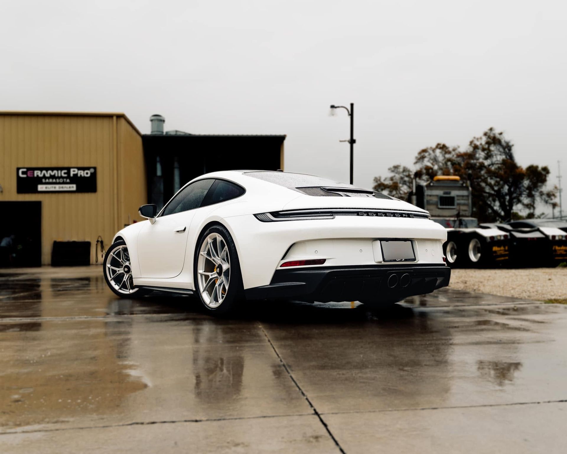 White sports car parked on a wet lot beside a tan building under an overcast sky