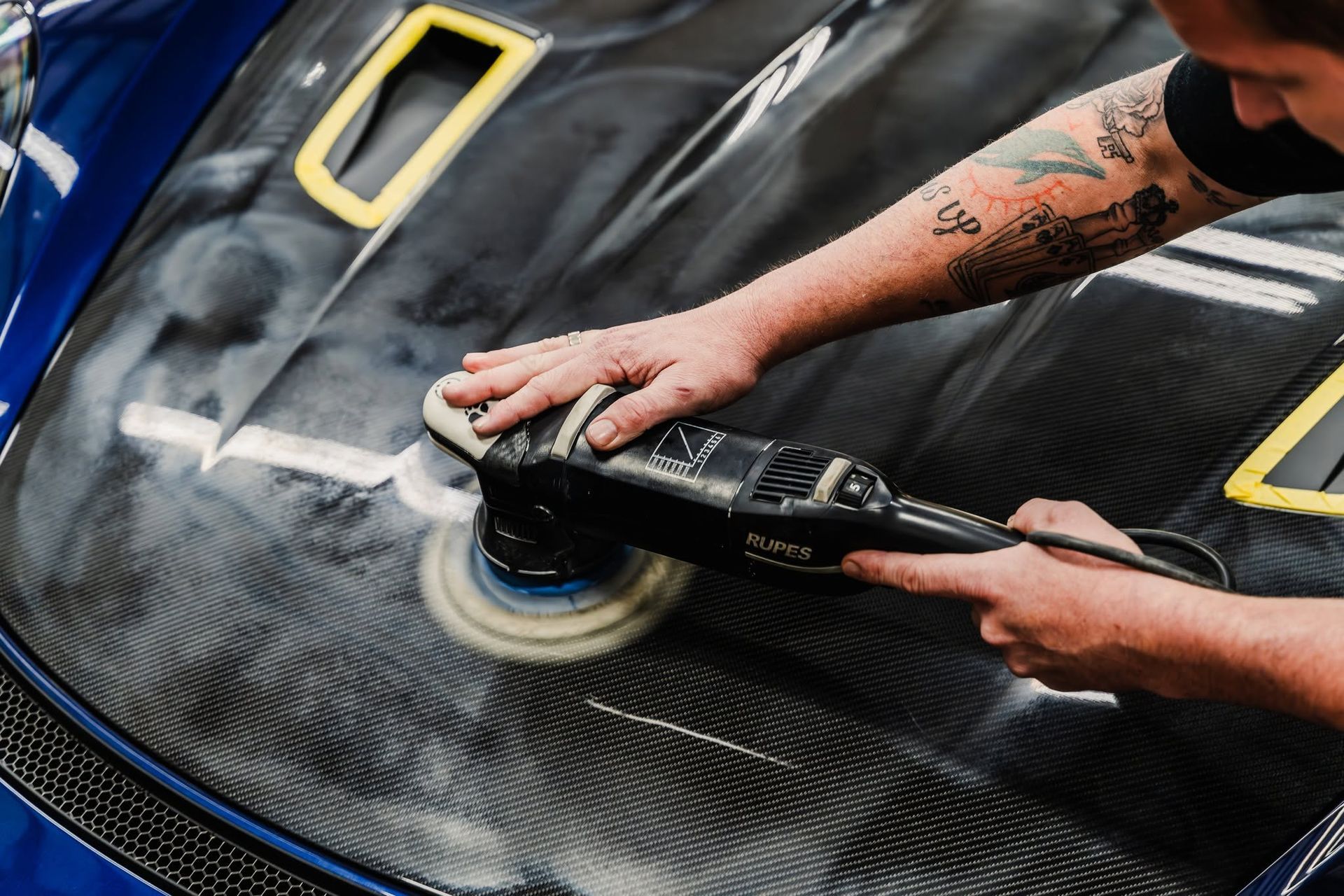 Person polishing a black sports car with a buffer, creating a glossy swirl on the hood