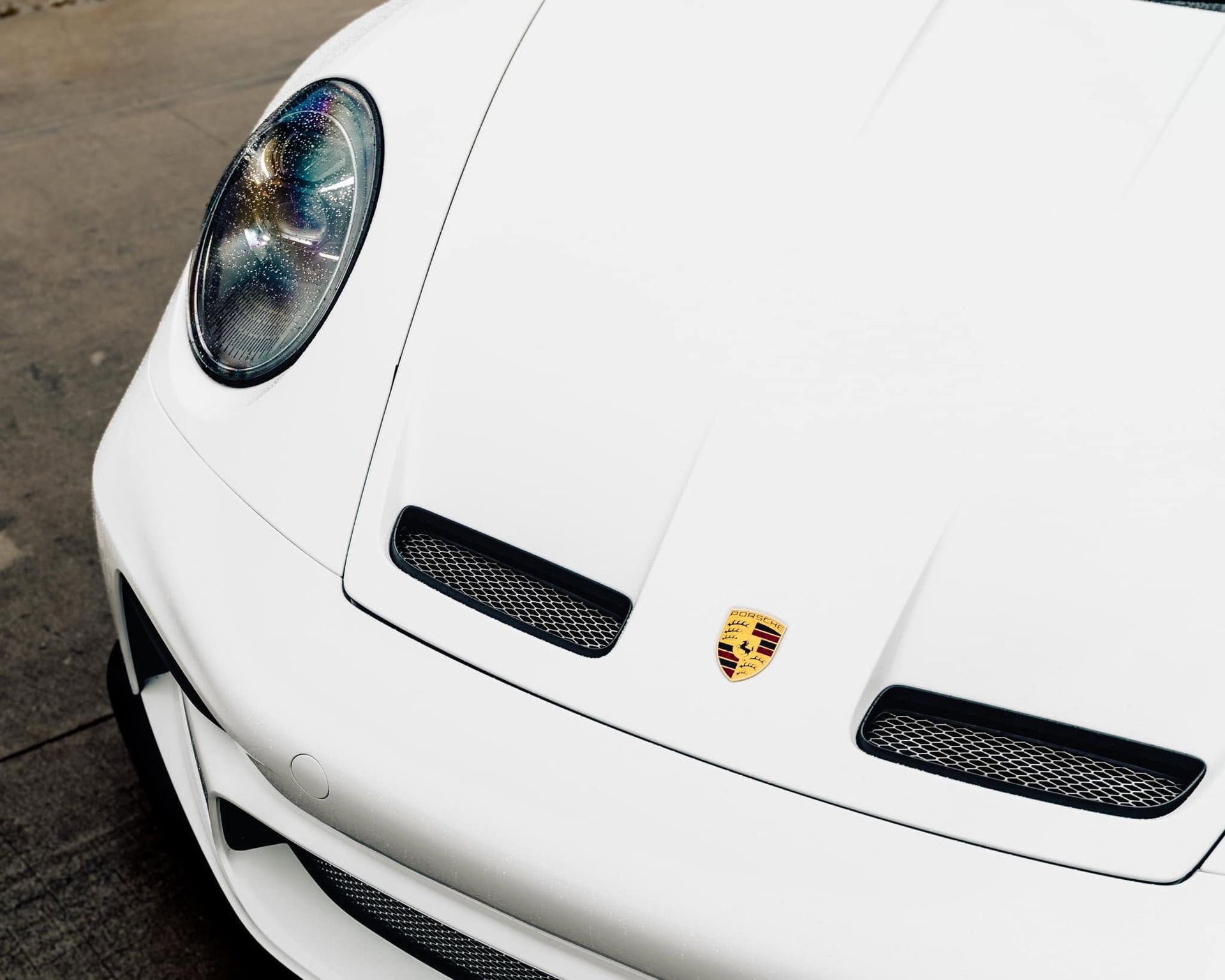 White Porsche sports car front end with headlight and hood vents, viewed from above-left