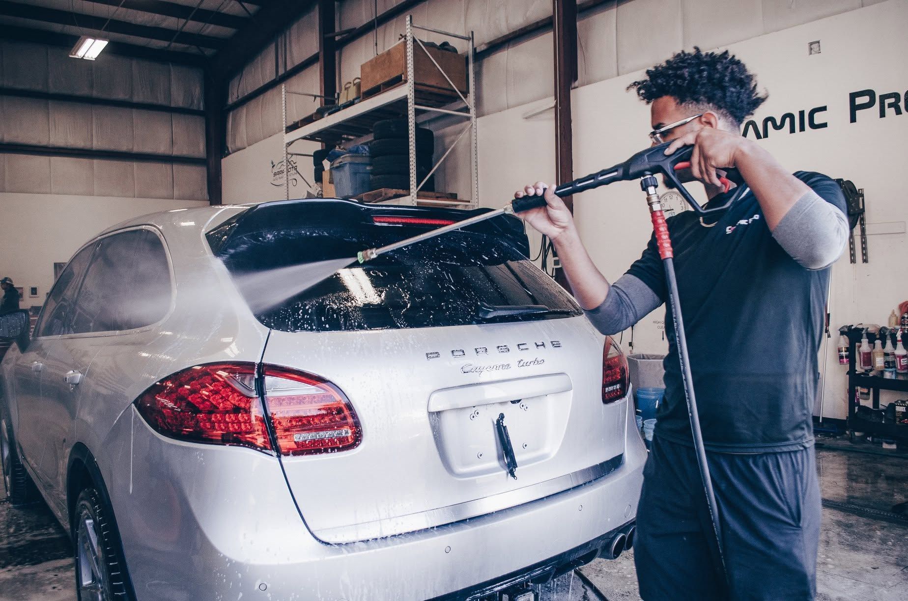 Man washing a silver SUV with a pressure washer in an auto detailing shop