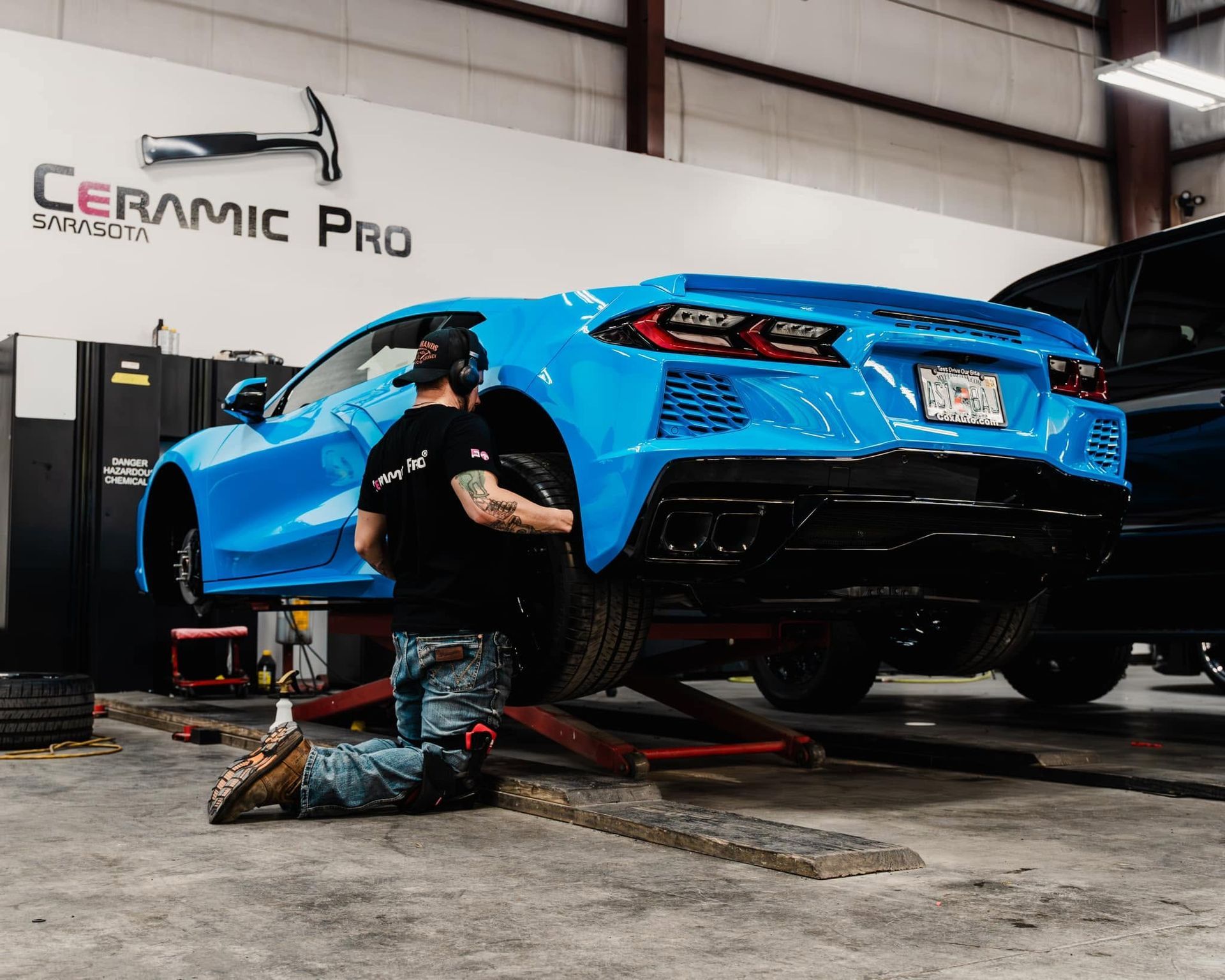 Blue sports car lifted in a garage while a mechanic works underneath near a Ceramic Pro sign