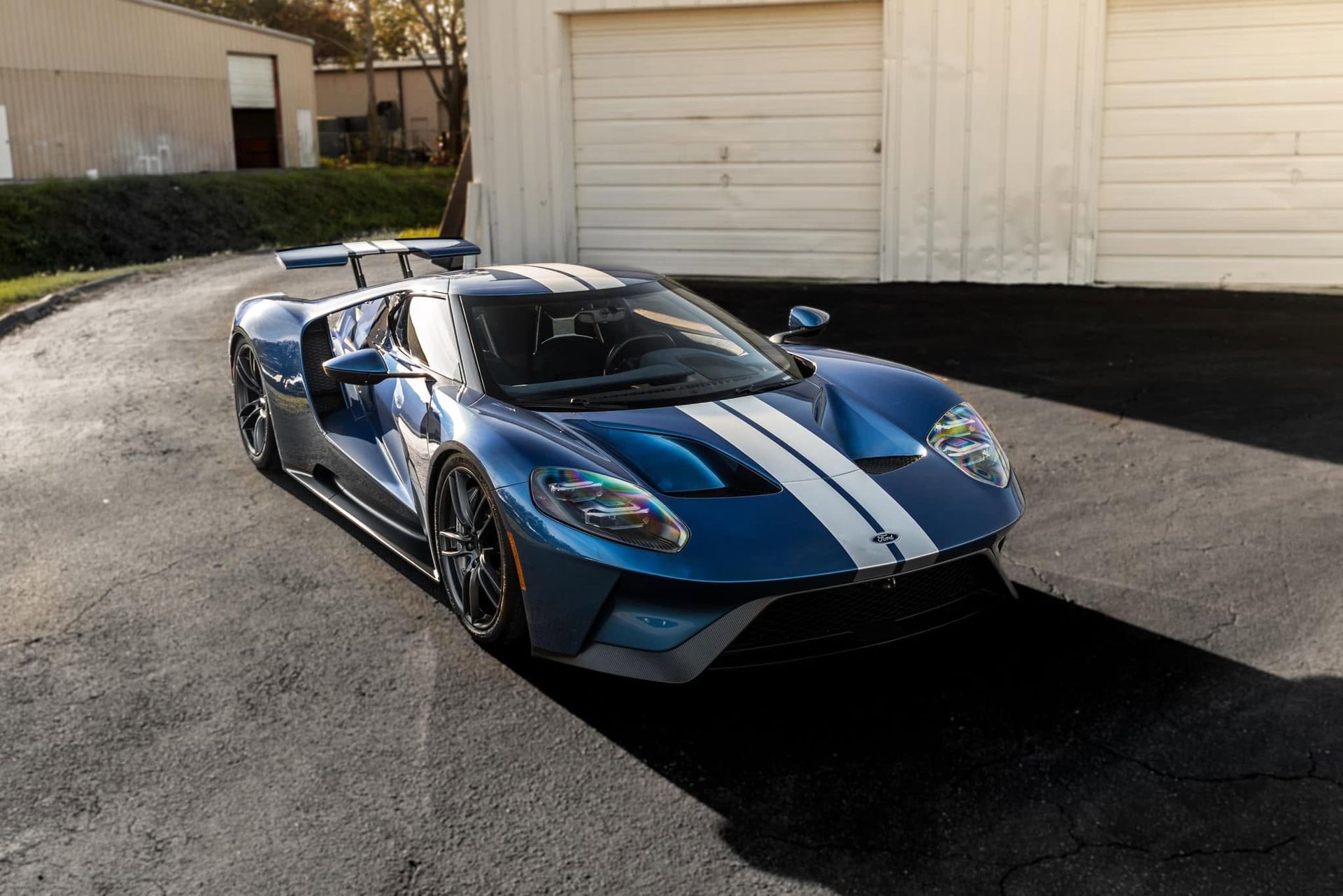 Blue Ford GT sports car with white racing stripes parked on asphalt in front of a garage.