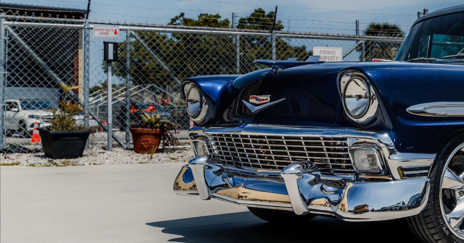 Front view of a shiny blue classic car parked by a chain-link fence in sunlight.