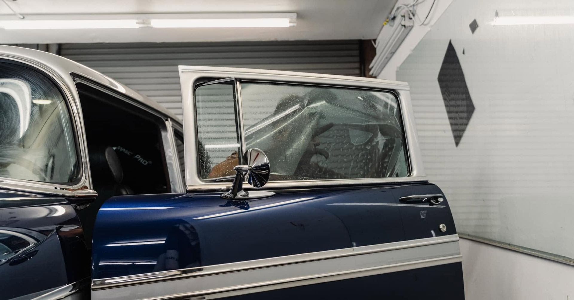 Dark blue vintage car in a garage, door open, with a white wall and overhead lights behind it