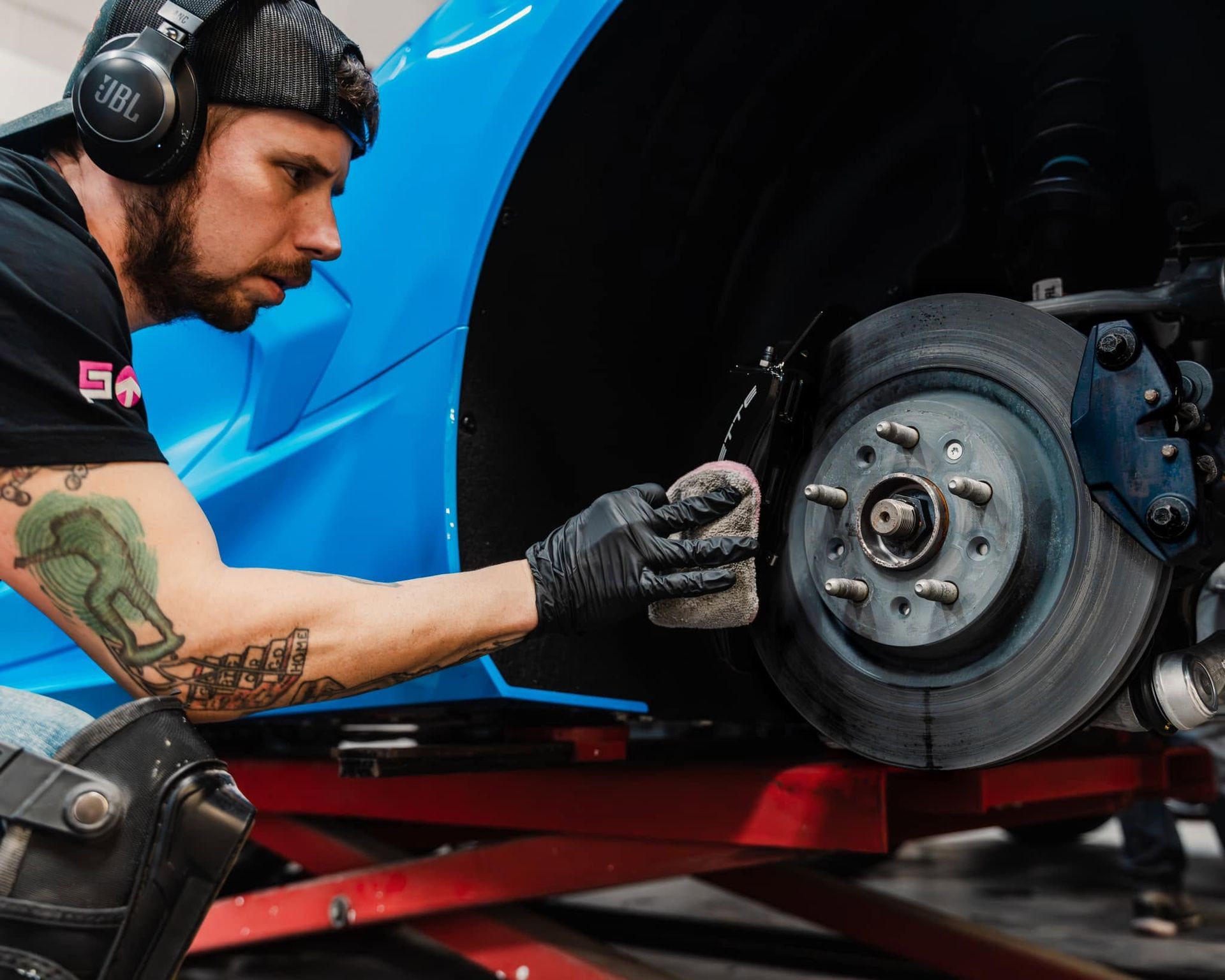 Mechanic in headphones works on a car brake assembly on a lift, wearing gloves and a black cap