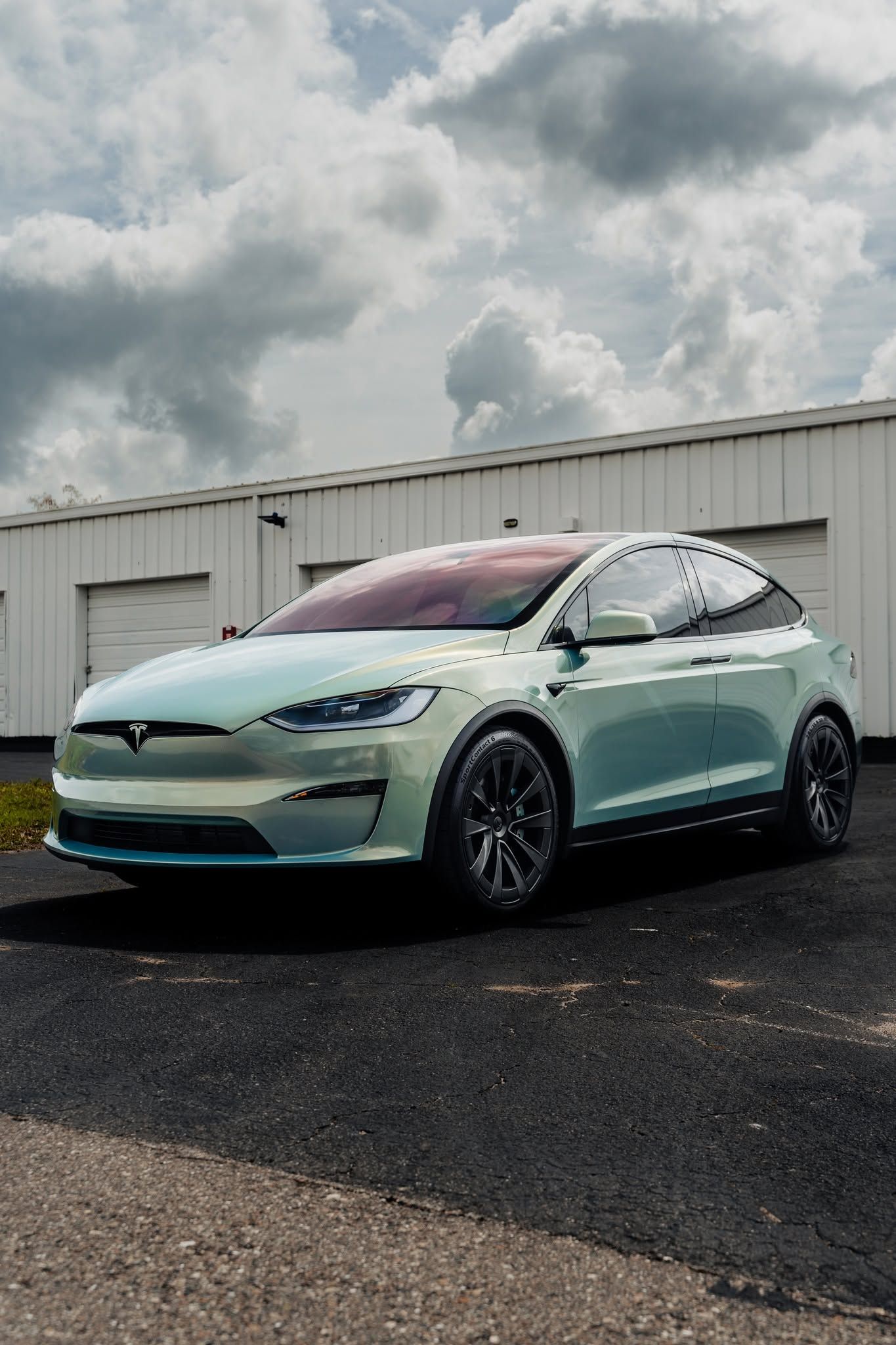Mint-green Tesla parked on wet pavement beside a white warehouse under cloudy skies