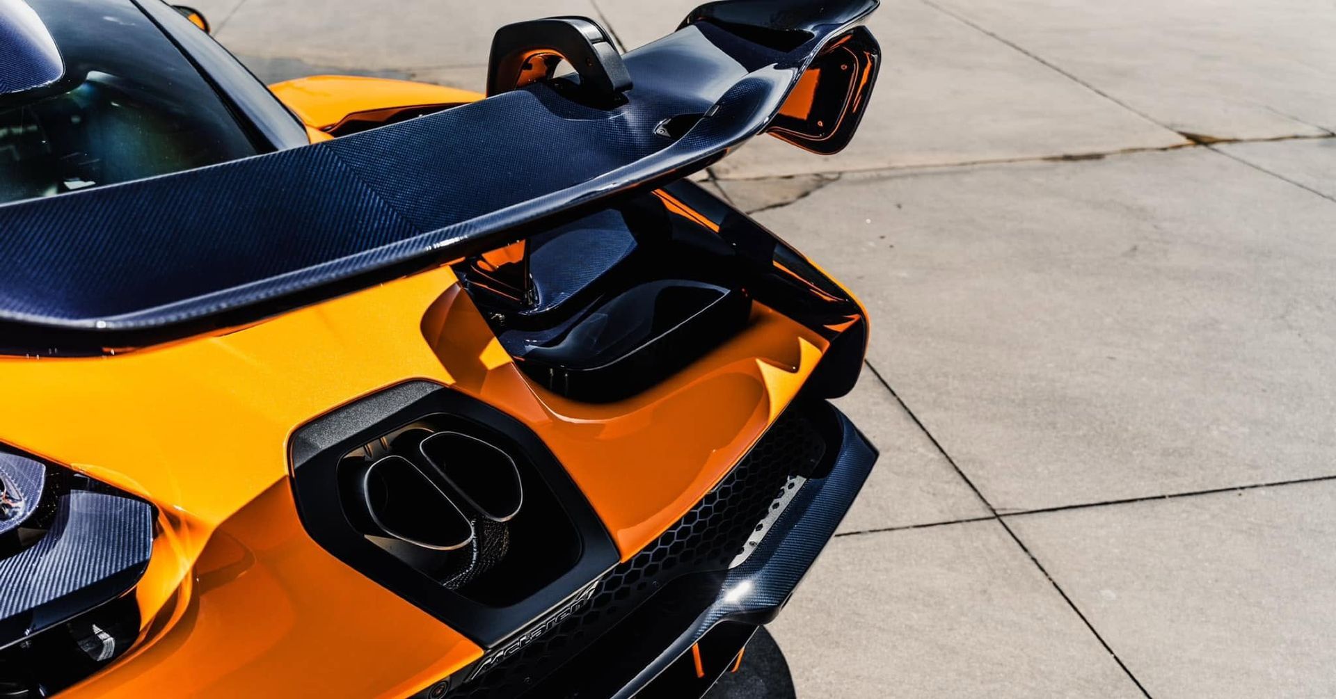 Orange sports car rear with black spoiler and exhausts, parked on concrete pavement