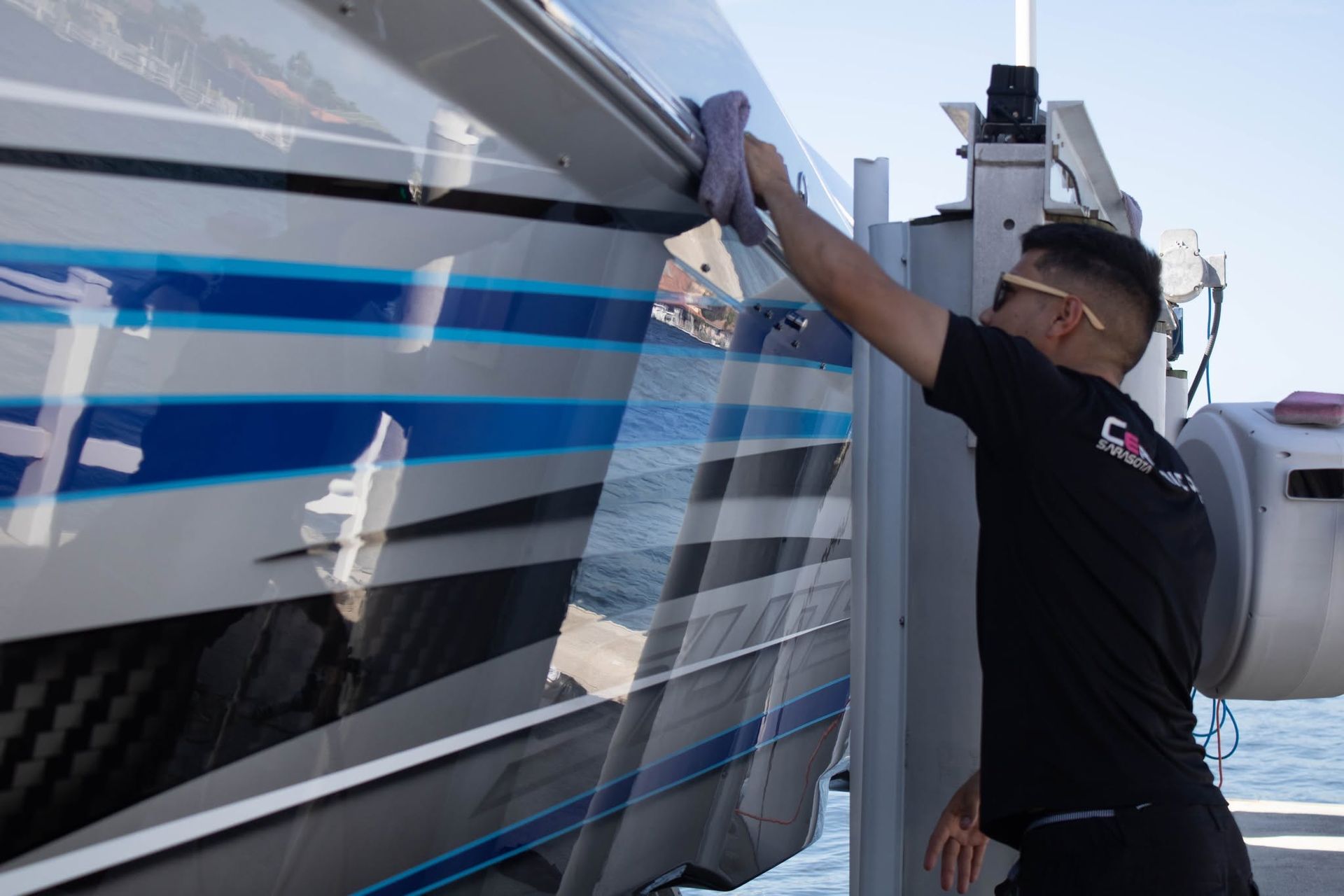 Man in black shirt wipes the side of a white boat with blue stripes at a marina