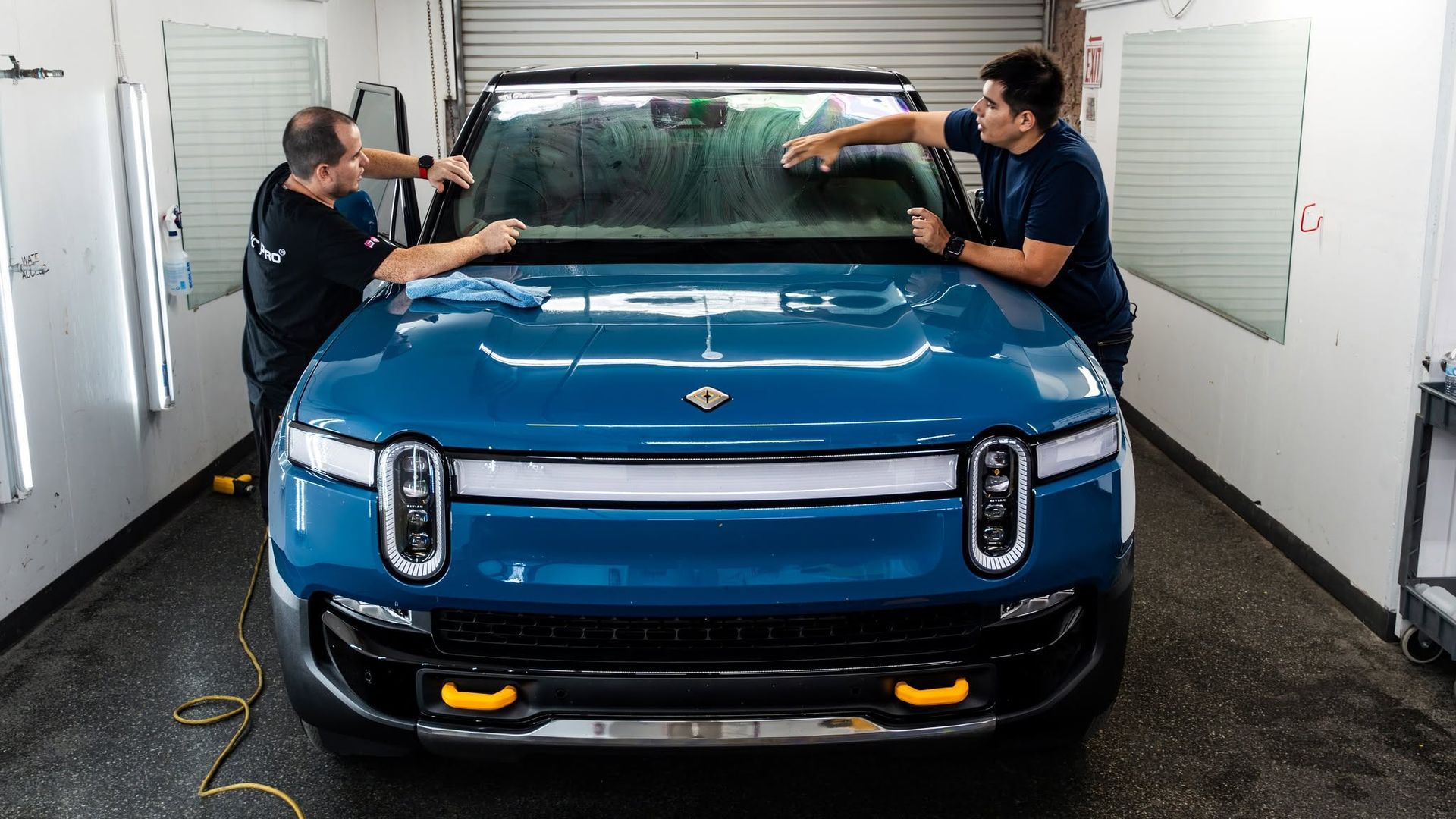 Two people installing a windshield on a blue pickup truck in a workshop