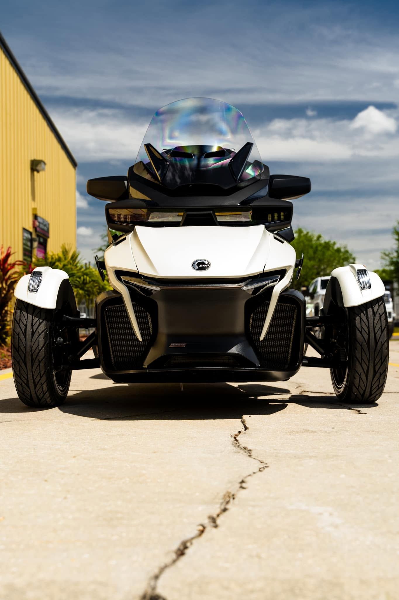 Three-wheeled white and black roadster parked on a driveway beside a yellow building under a blue sky