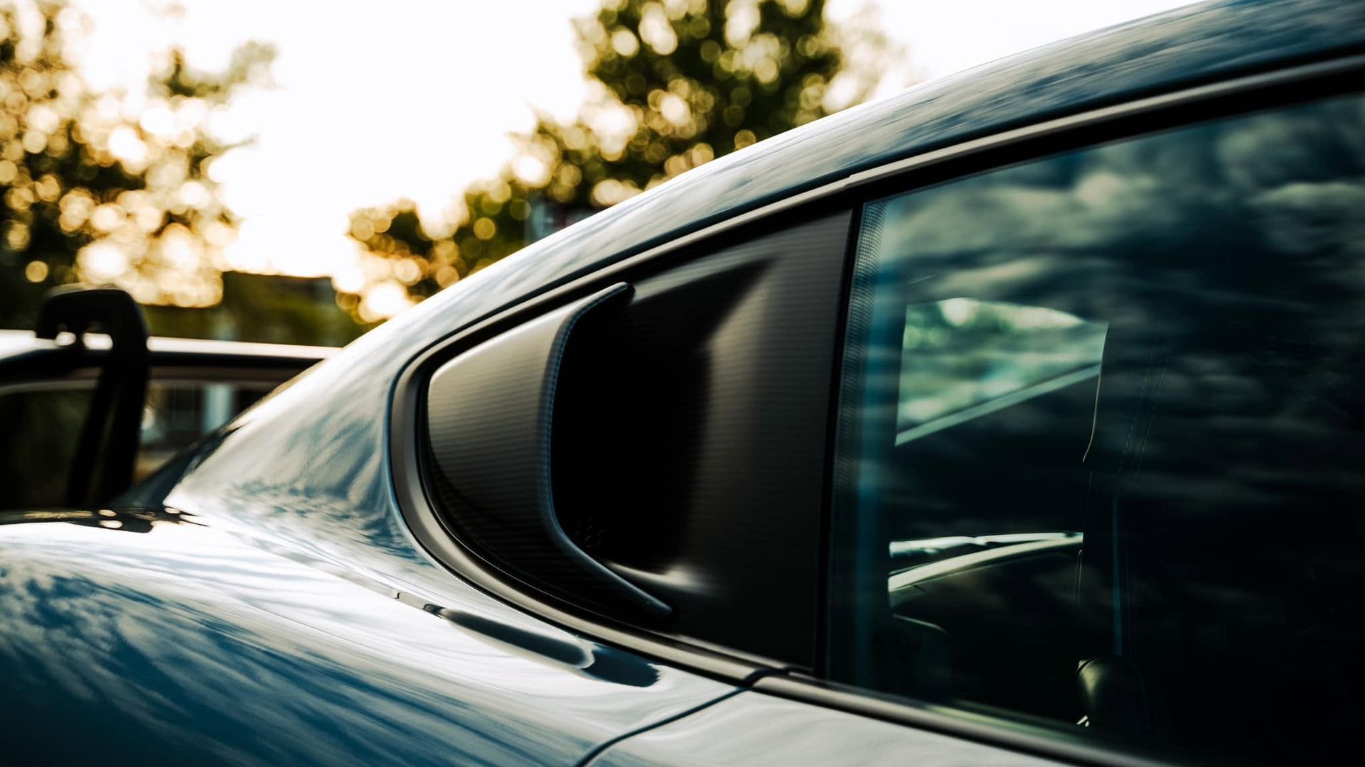 Close-up of a blue car’s rear side window and roofline at sunset, with trees reflected in the glass