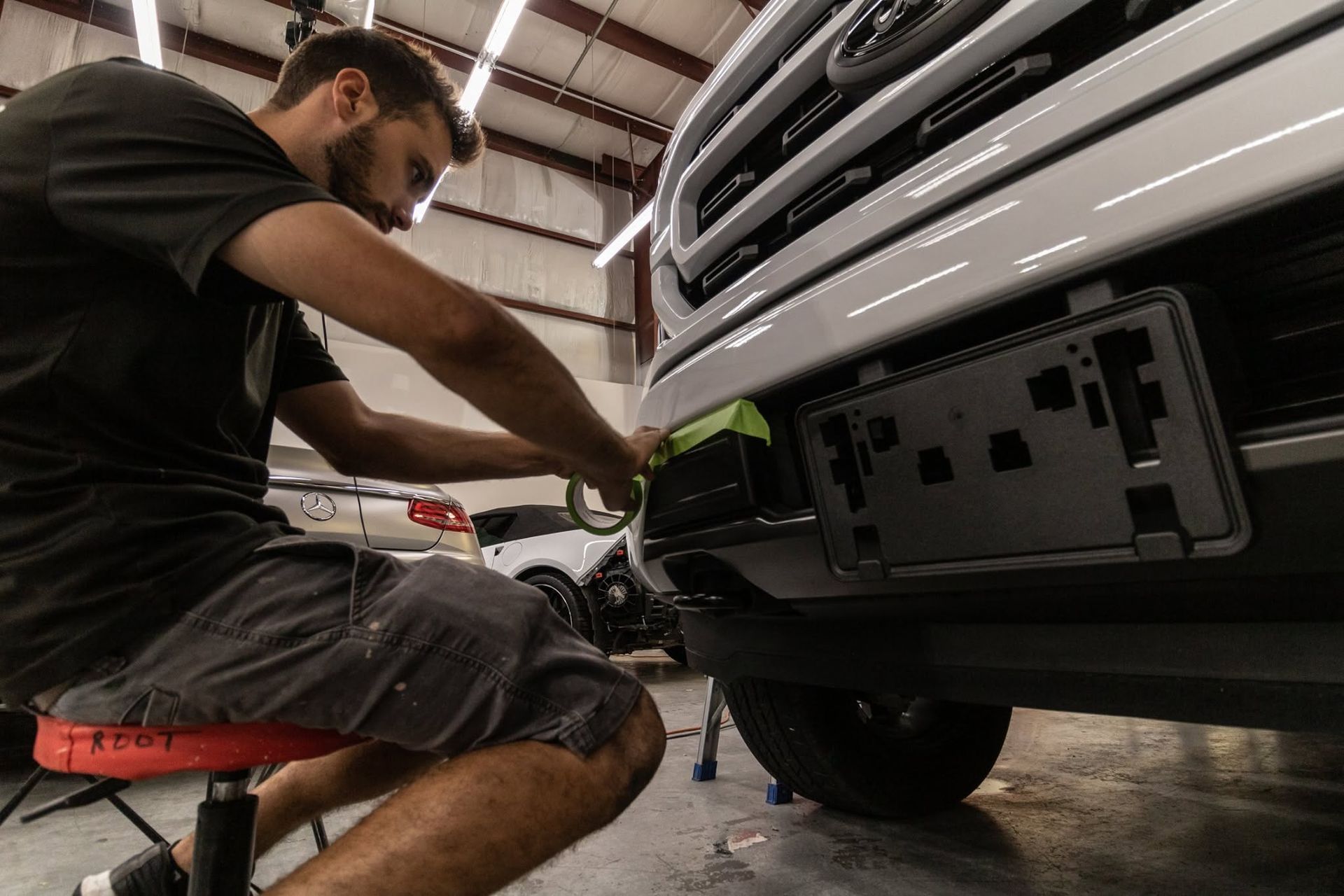 Mechanic kneeling beside a silver vehicle, working under the front bumper in a garage
