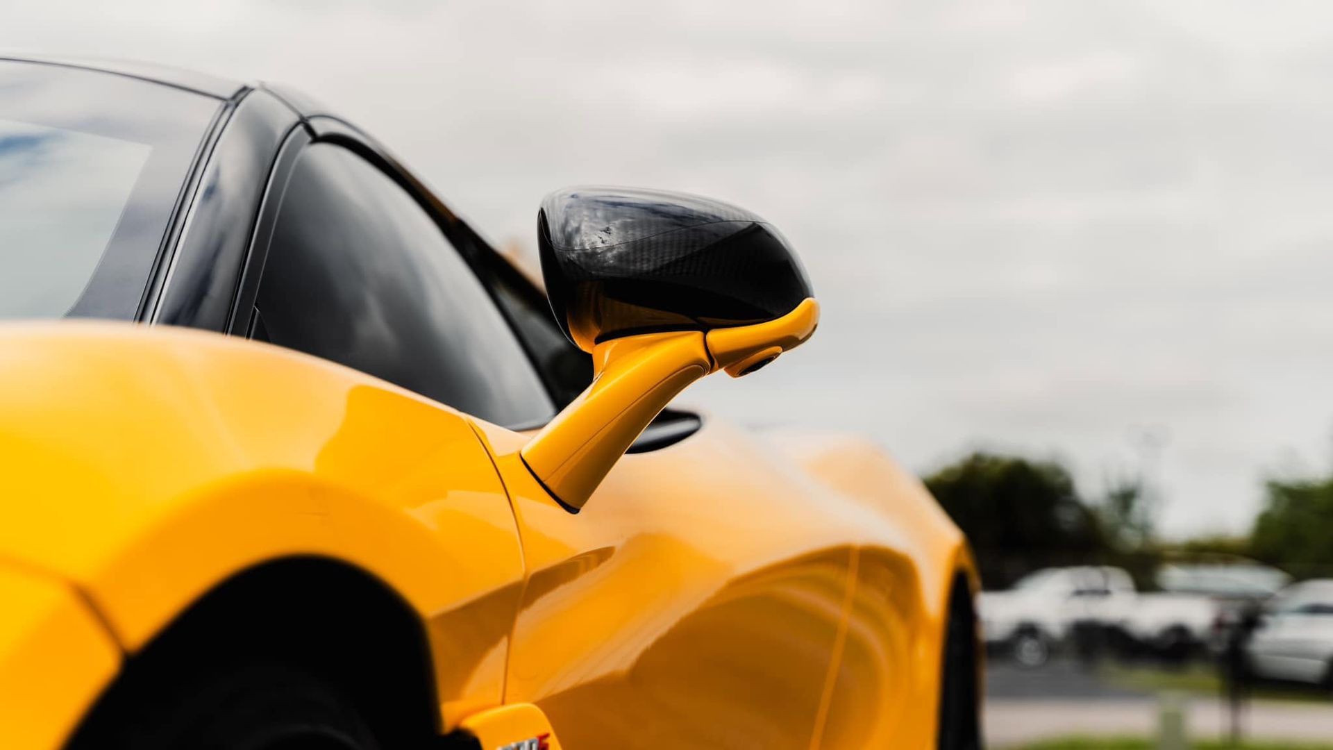 Close-up of a yellow sports car with a black side mirror on a road under a cloudy sky