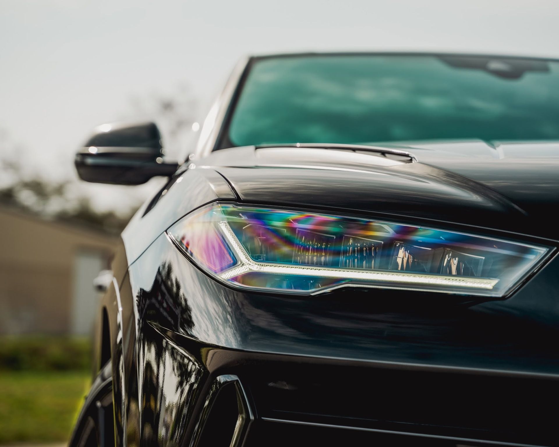 Close-up of a black car’s front headlight with a rainbow reflection outdoors