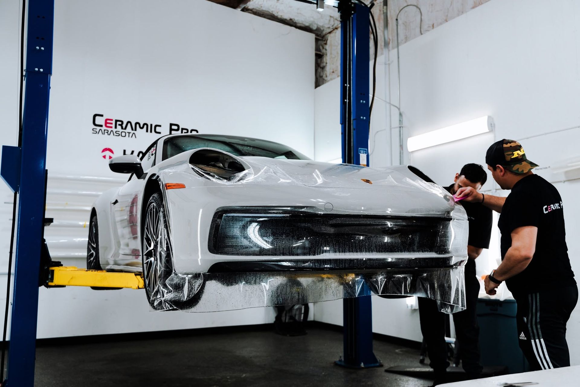White sports car being wrapped in a ceramic coating shop, with two workers on the right.