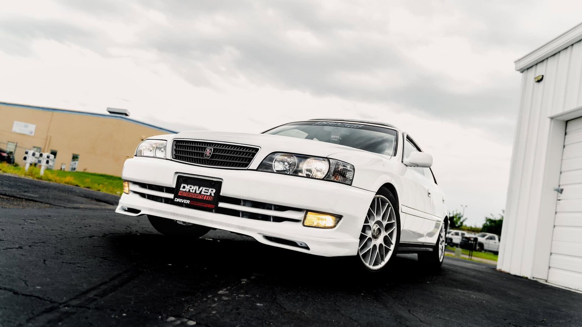 White sports car parked on wet pavement beside a white building on an overcast day