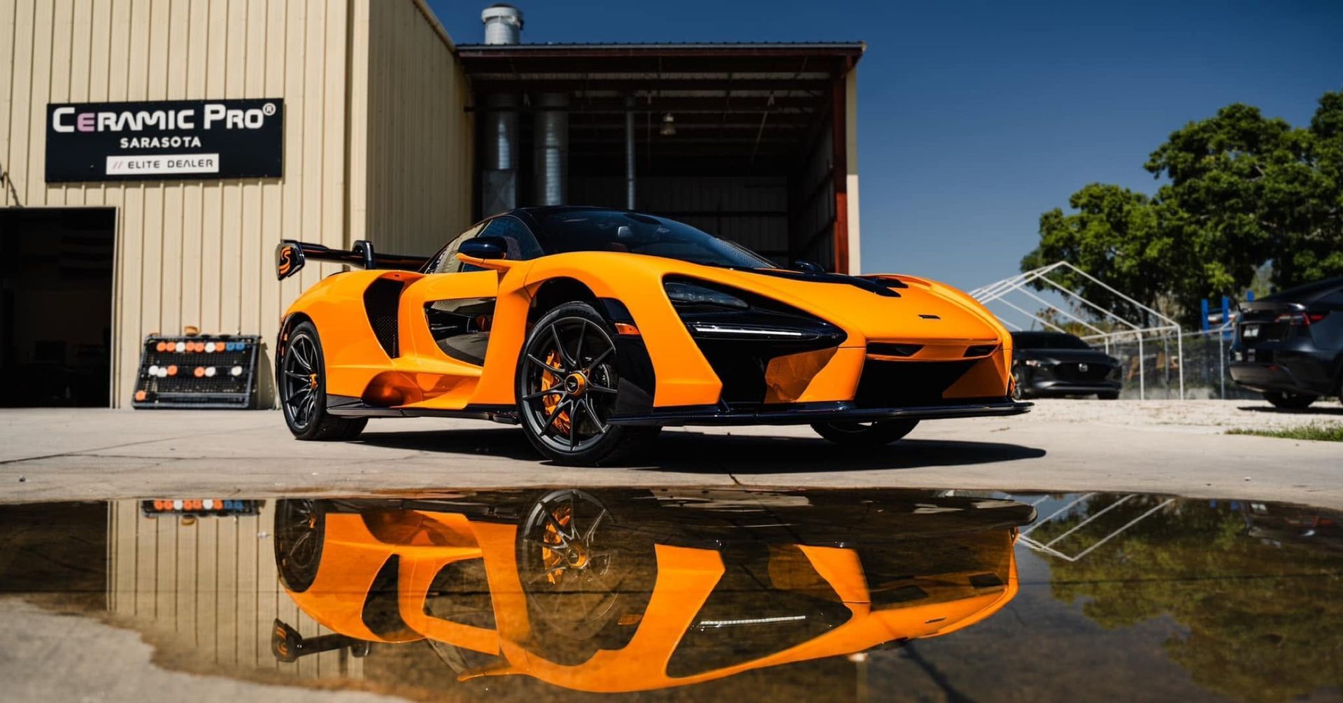 Orange sports car reflected in a puddle outside Ceramic Pro shop under a bright sky