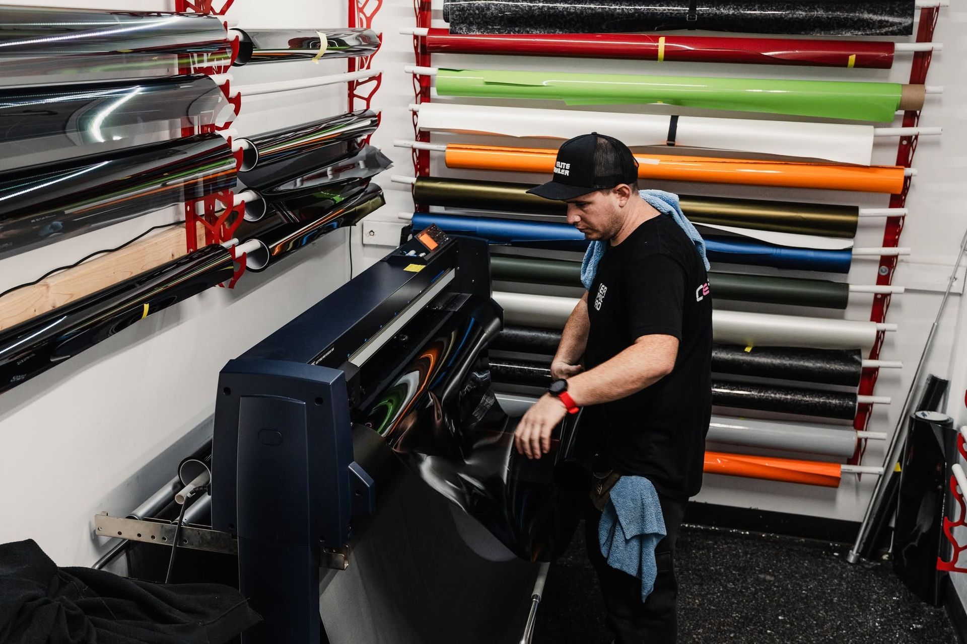 Person working with a large-format printer in a print shop, surrounded by rolls of colorful vinyl.
