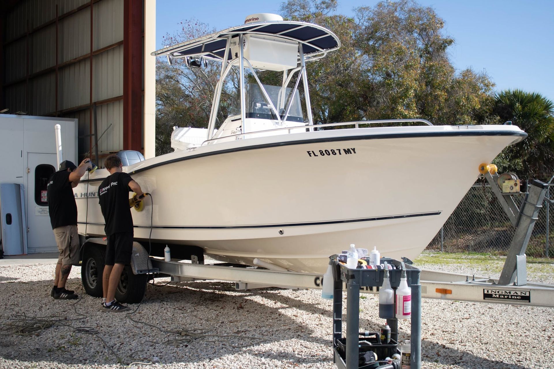 Two people inspect a white boat on a trailer outside a metal building.