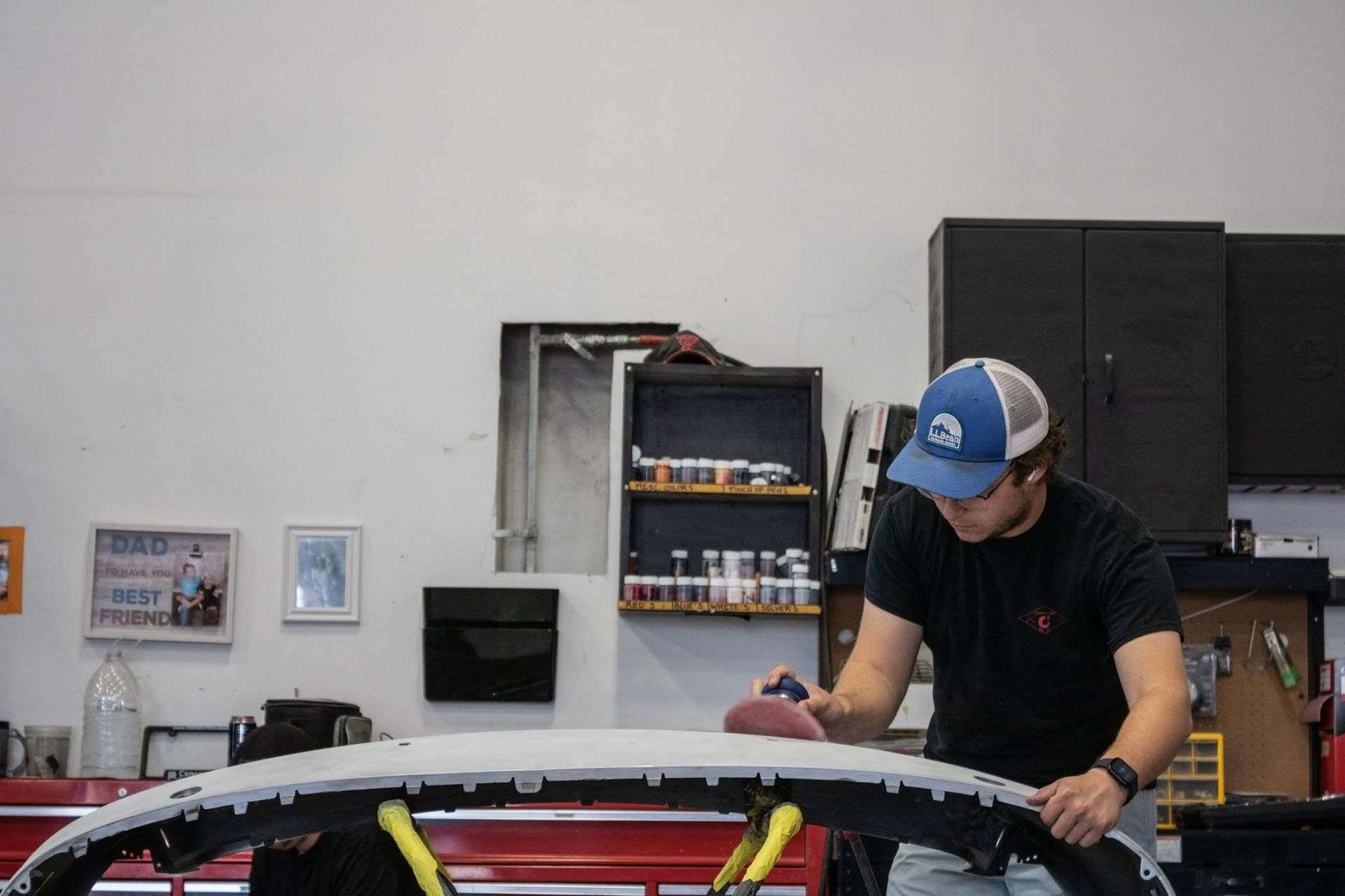 Person in a blue cap works on a car in a garage, leaning over the roof with tools nearby.