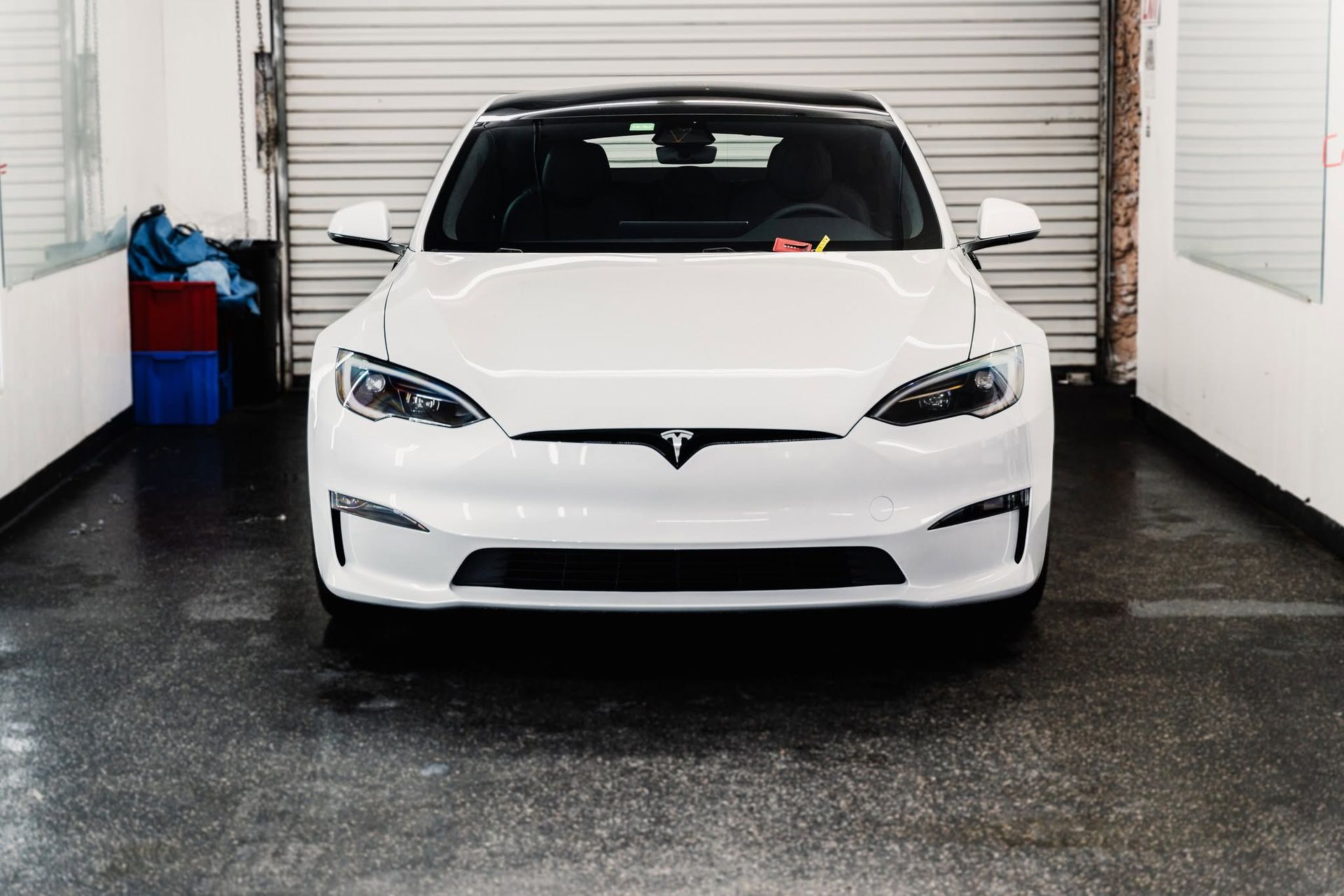 White Tesla sedan parked in a garage, facing forward with the hood badge visible.