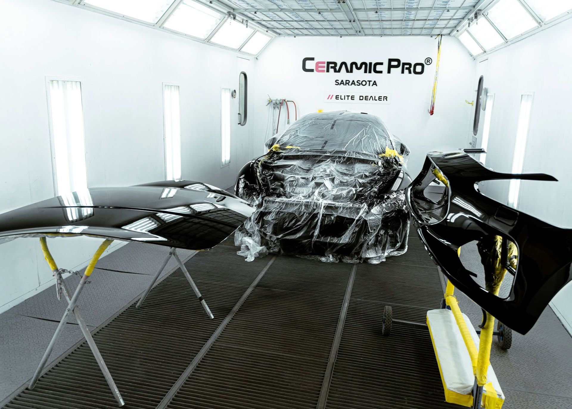 Sports car wrapped in clear film inside a white Ceramic Pro detailing booth, with yellow work lights