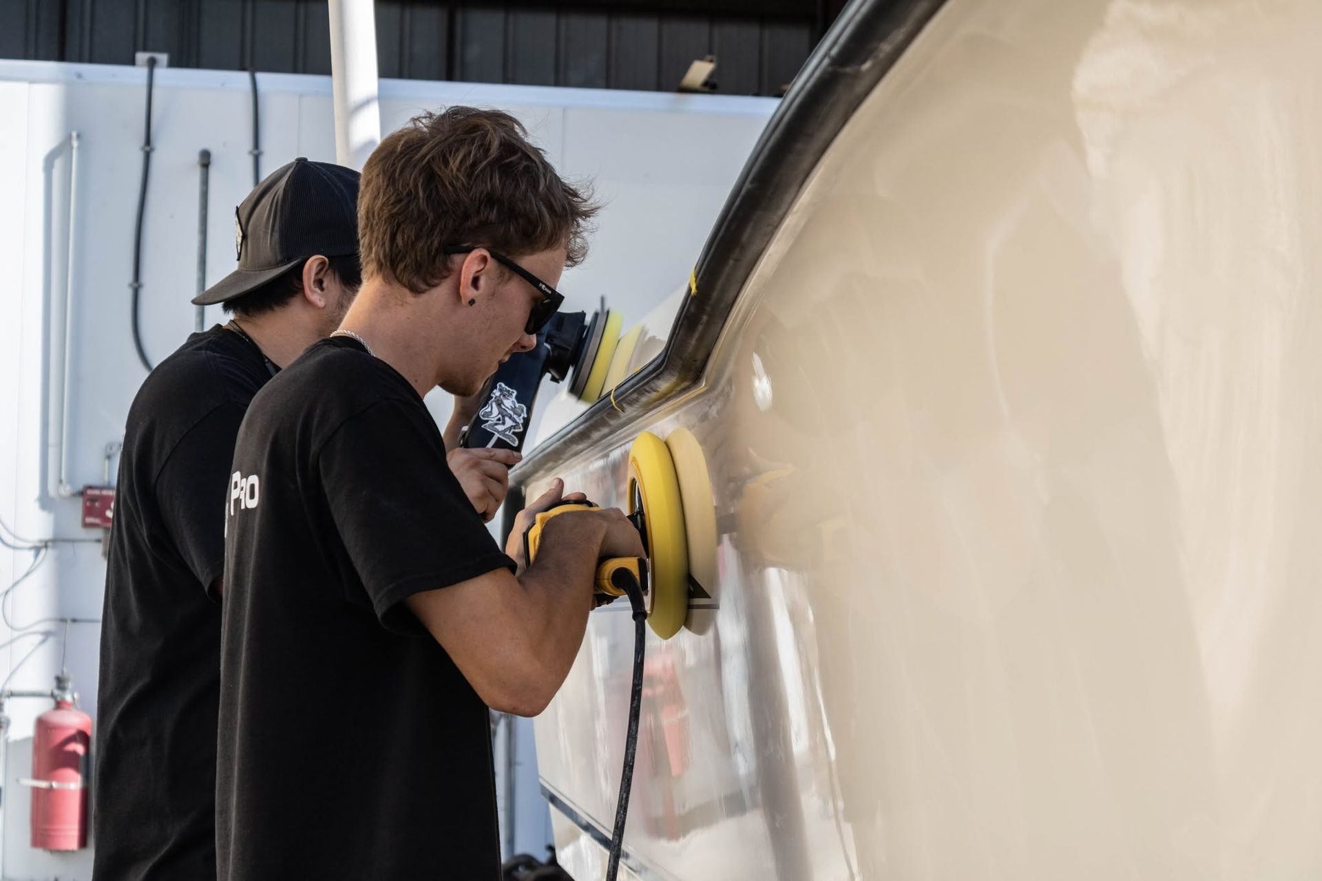 Two workers polish a beige vehicle panel with a buffer in a workshop.
