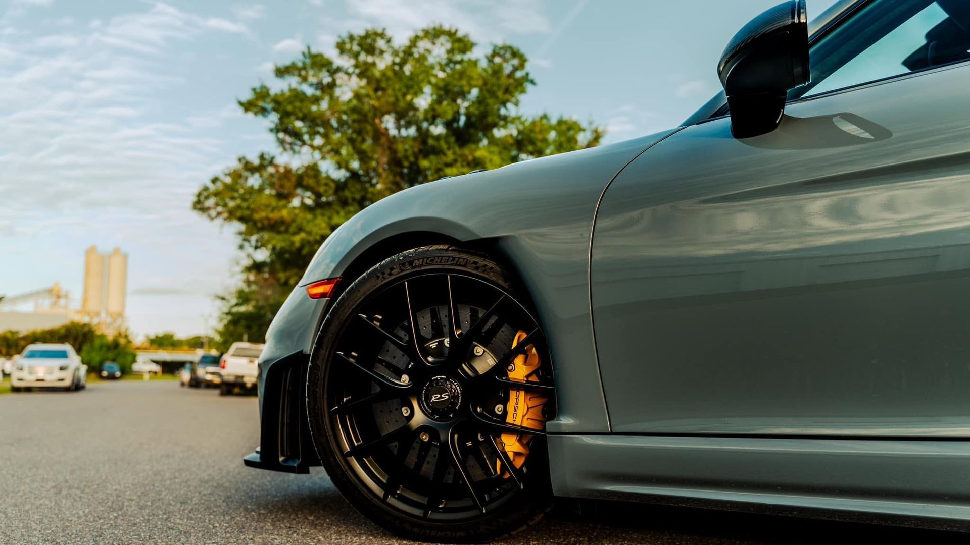 Close-up of a gray sports car’s black wheel with orange brake caliper, parked on a road near trees at sunset