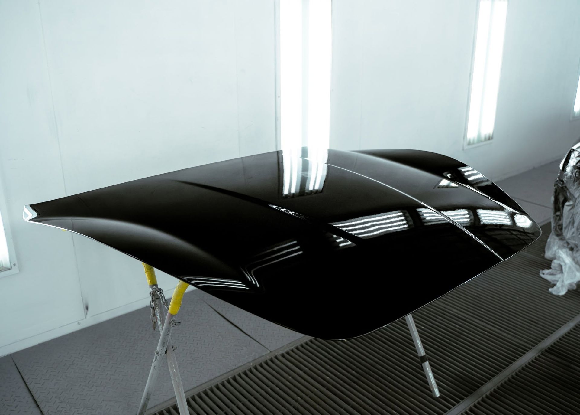 Glossy black car hood in a spray booth, propped on stands under bright lights