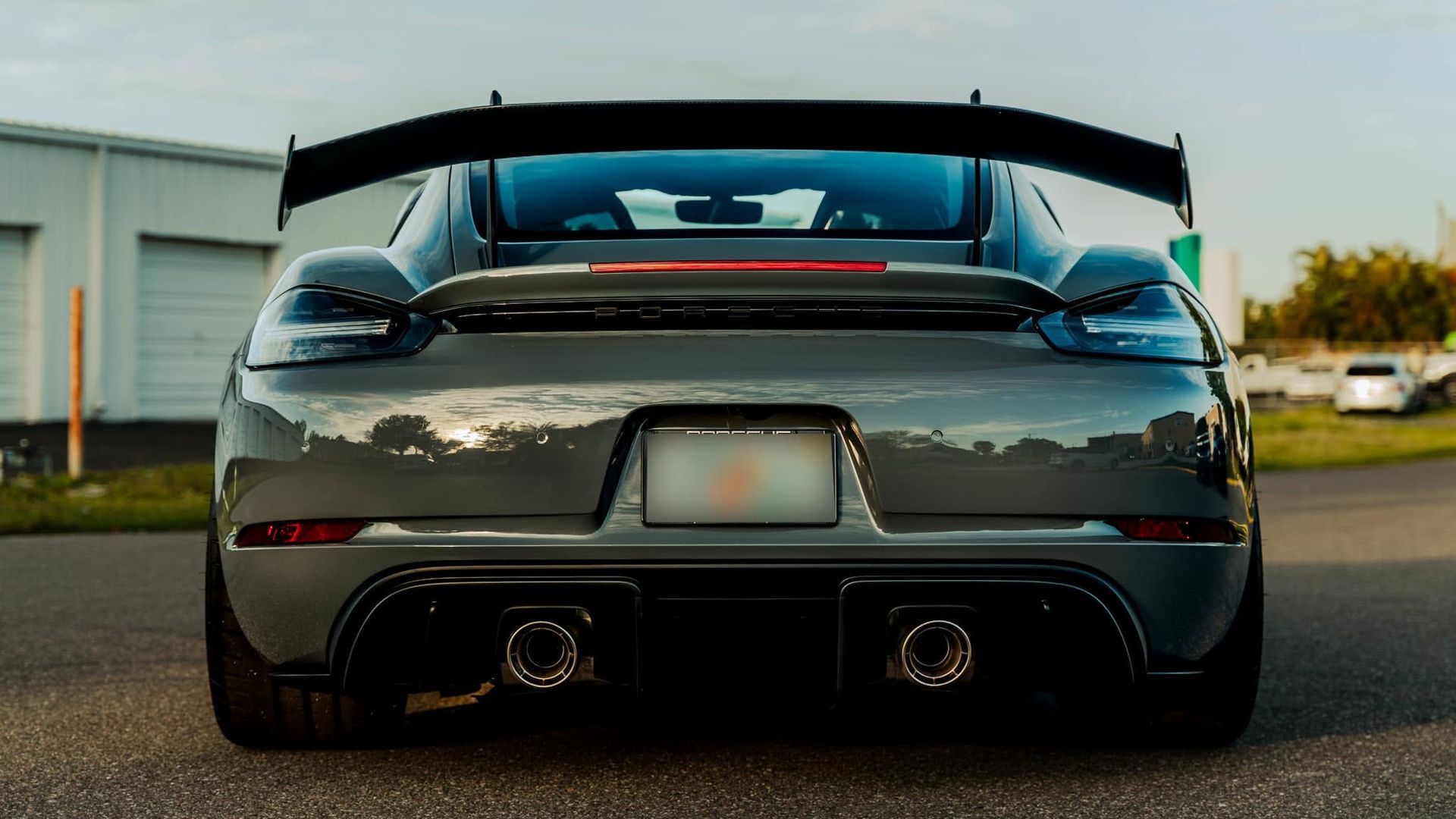 Rear view of a dark sports car with a large spoiler, parked on pavement at sunset.