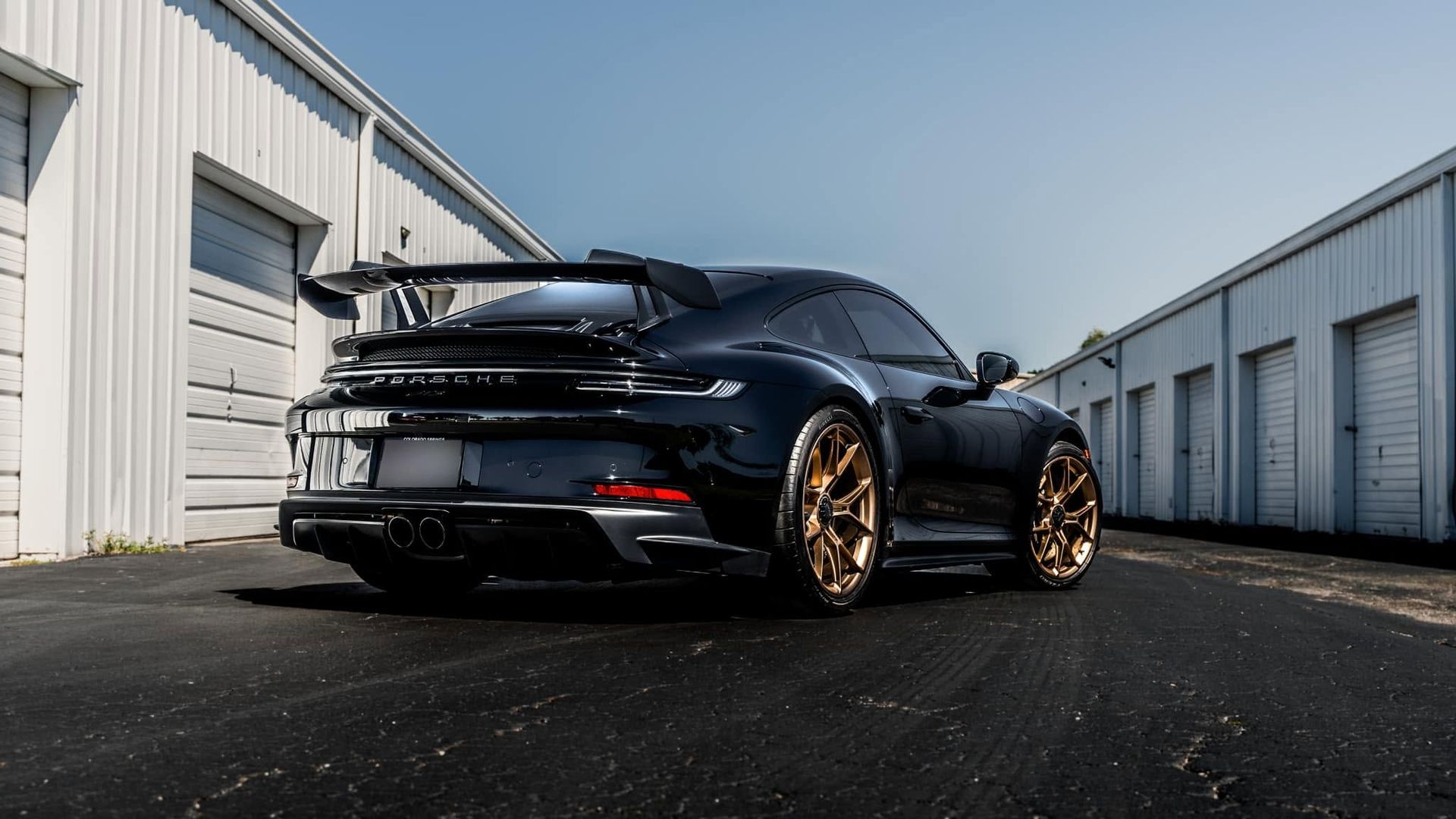 Black sports car with bronze wheels parked between white warehouse buildings on an industrial lot.