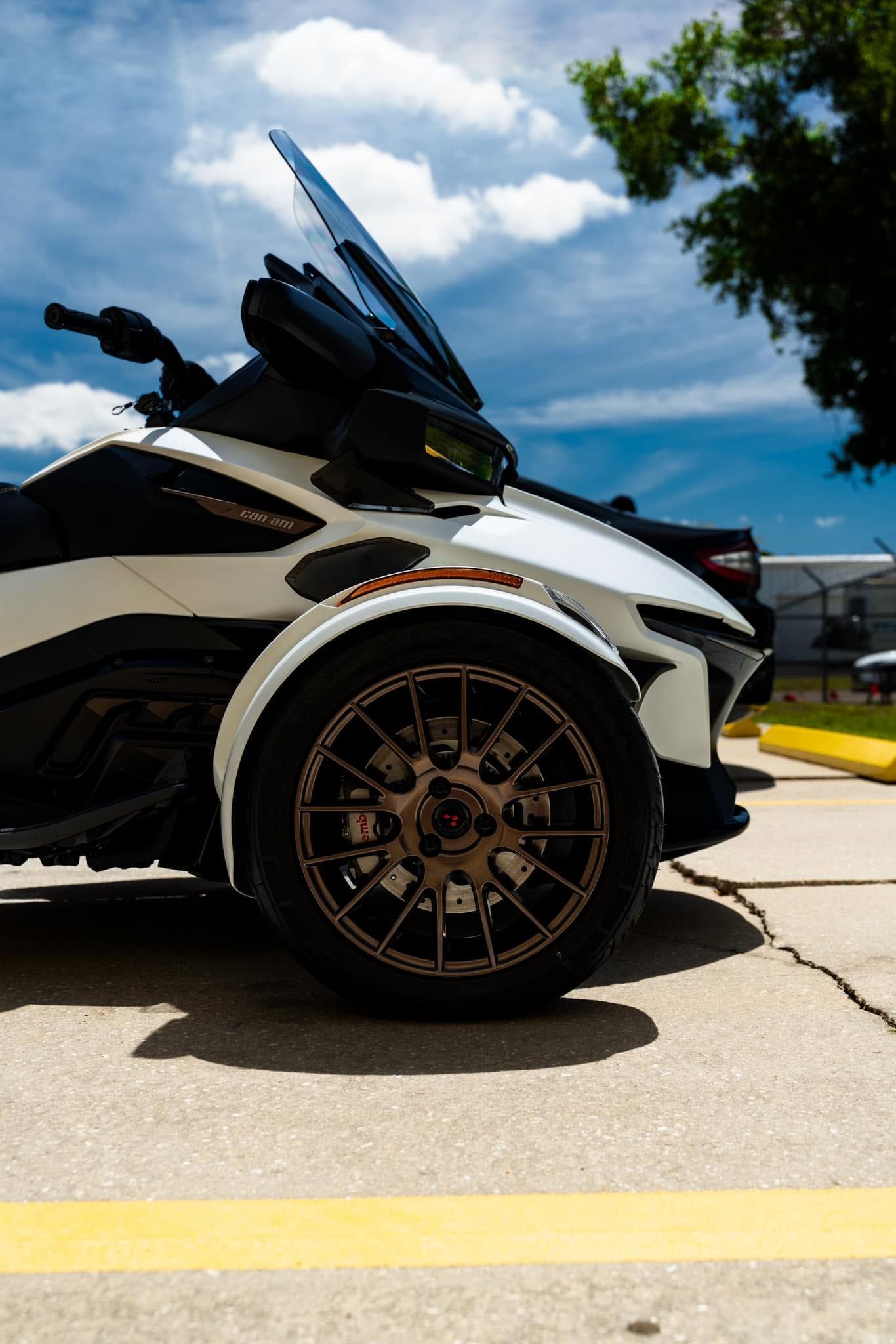 Customized white and black sports car with large bronze wheel, parked outdoors under a blue sky.
