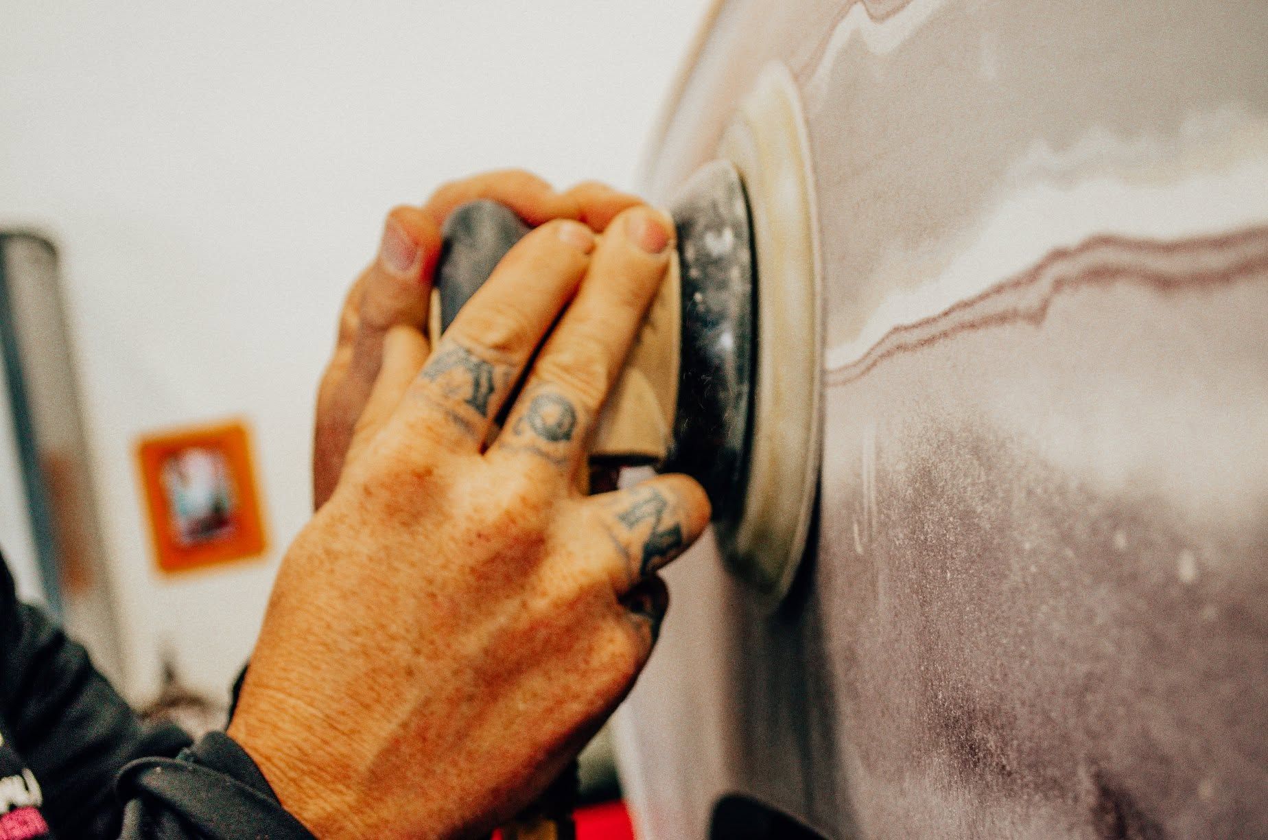 Hands sanding a car door with a round sander, creating dust on the metallic surface.