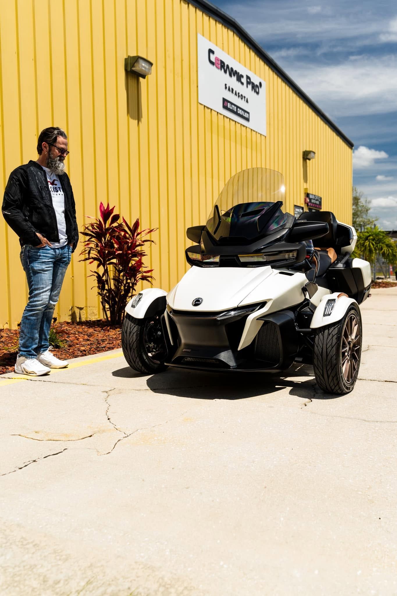 Man standing beside a white three-wheeled motorcycle outside a yellow building under a sunny sky