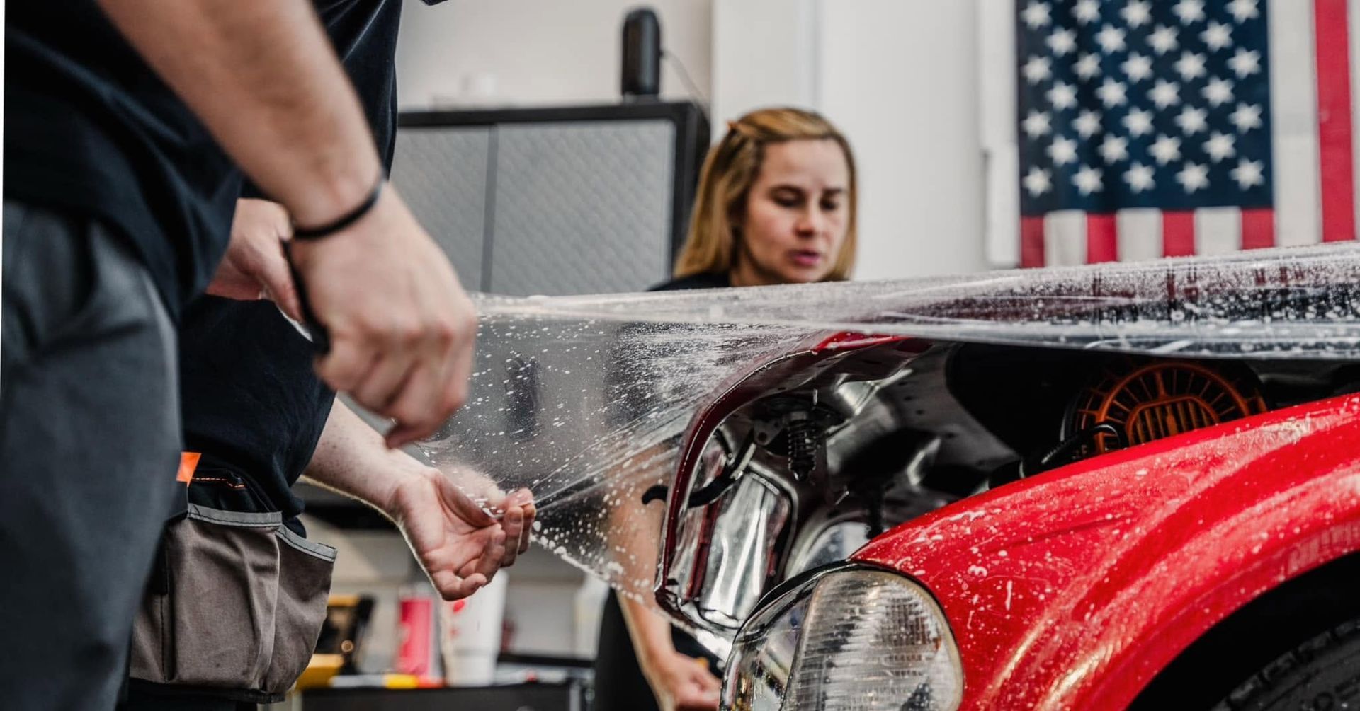 Person peeling clear protective film off a red sports car in a garage, with a blurred American flag in back.