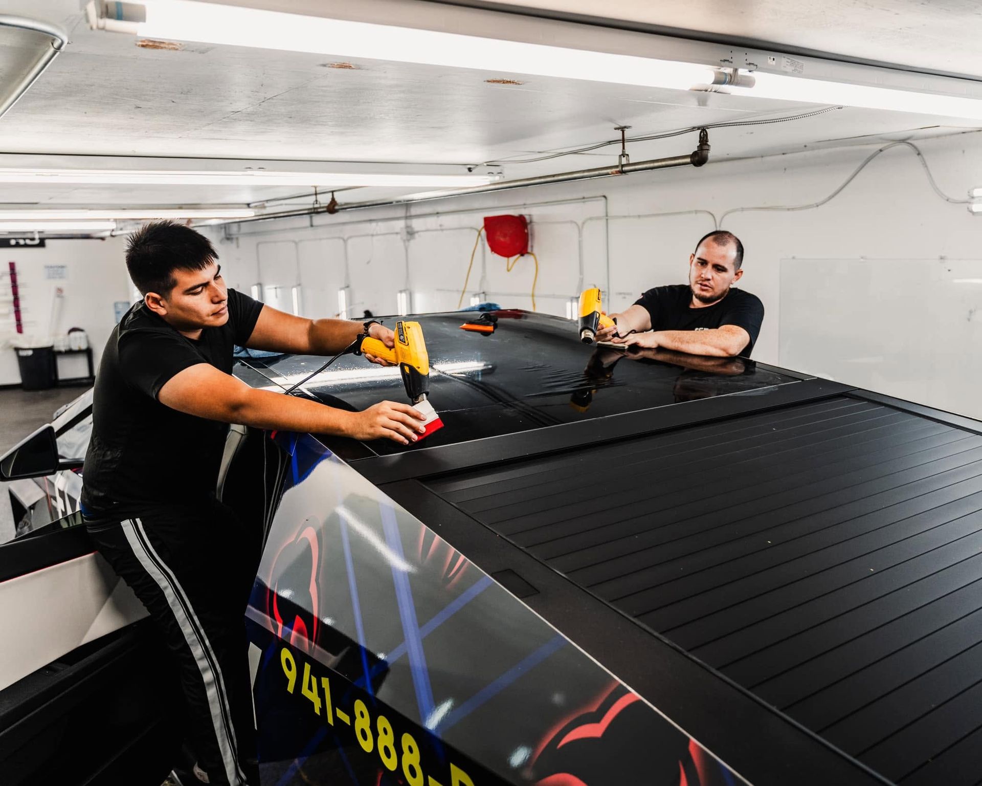 Two workers in black shirts securing a large black roof panel inside a workshop