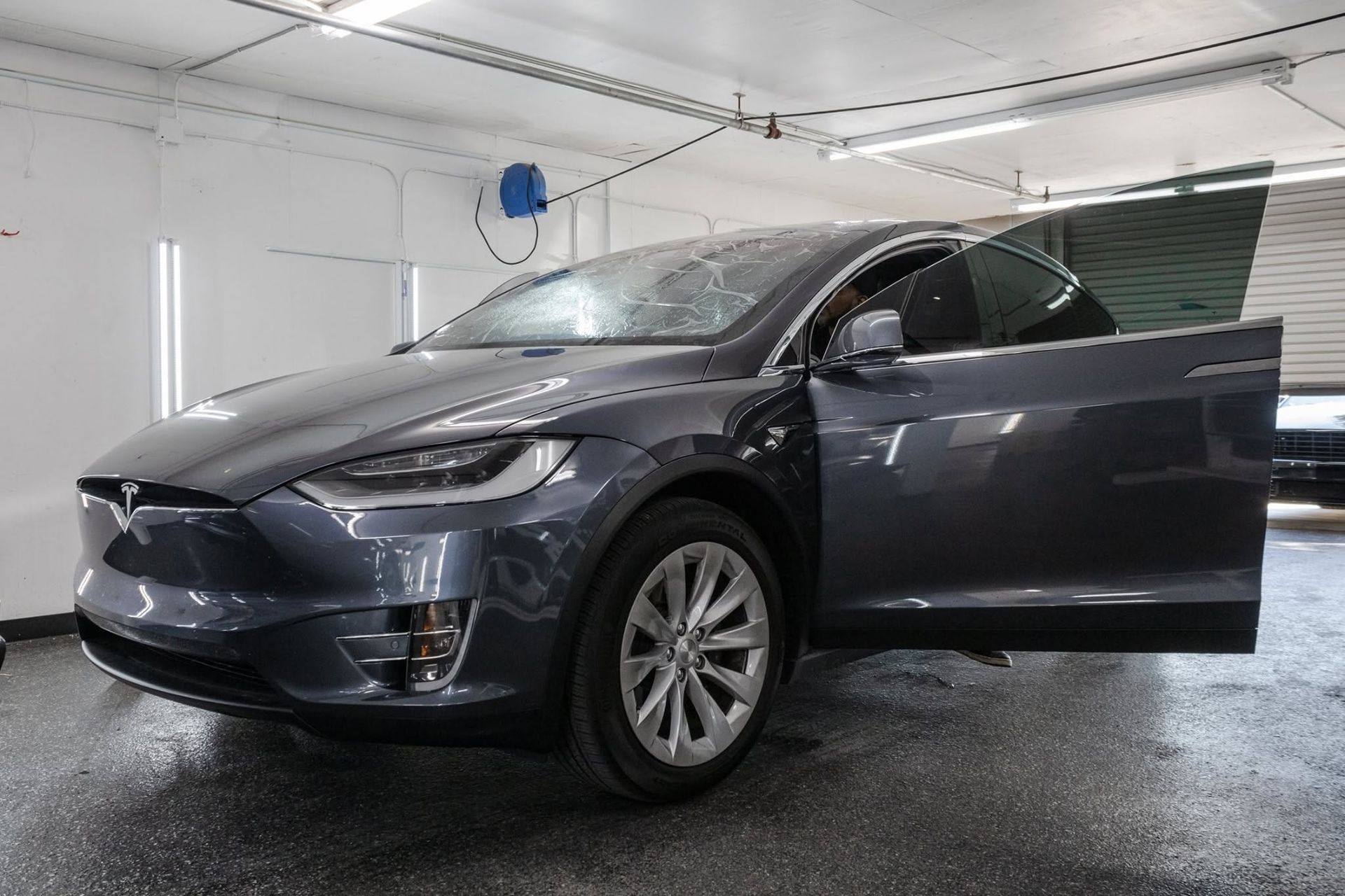Dark gray Tesla sedan in a garage, front door open and headlights on.