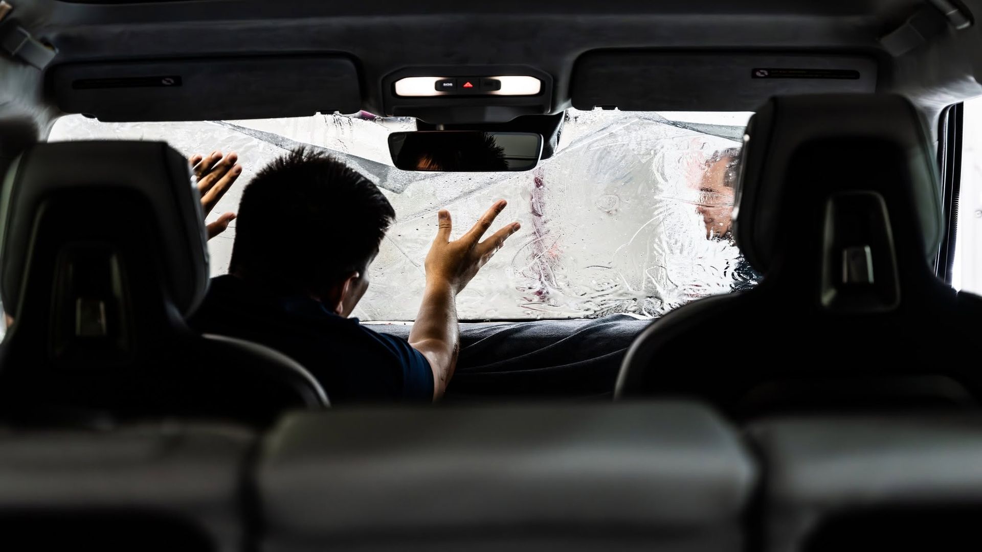 Backseat view of a person scraping snow and ice off a car windshield from inside the vehicle