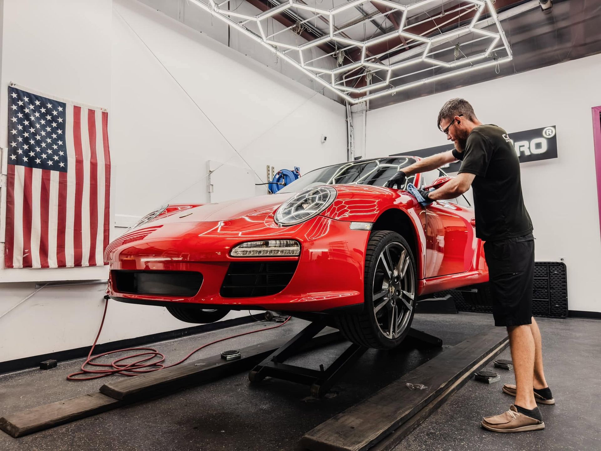 Red sports car on a dynamometer while a mechanic works on it in a garage with an American flag.