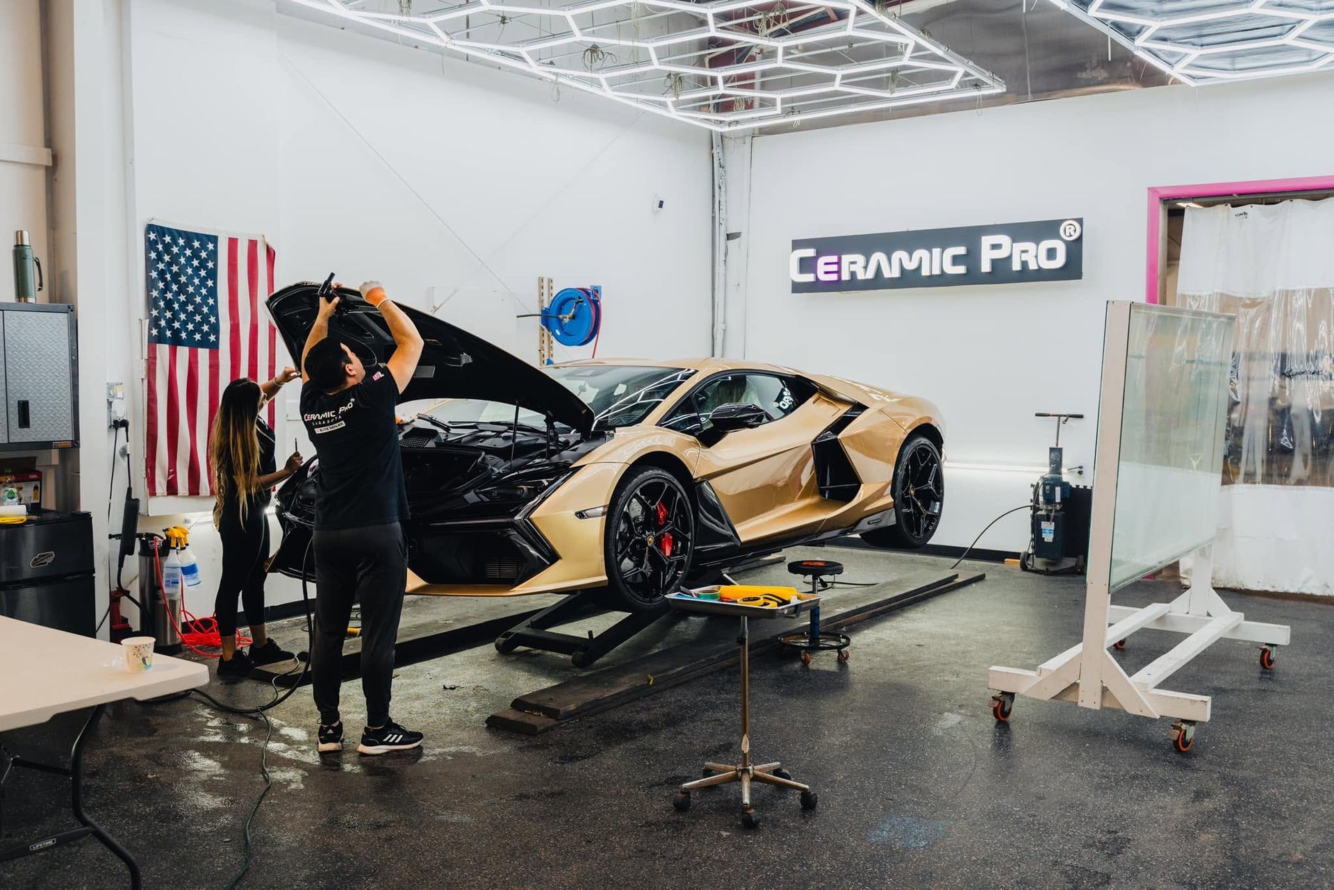 Detailer wrapping a gold sports car in a bright auto shop with a “Ceramic Pro” sign.
