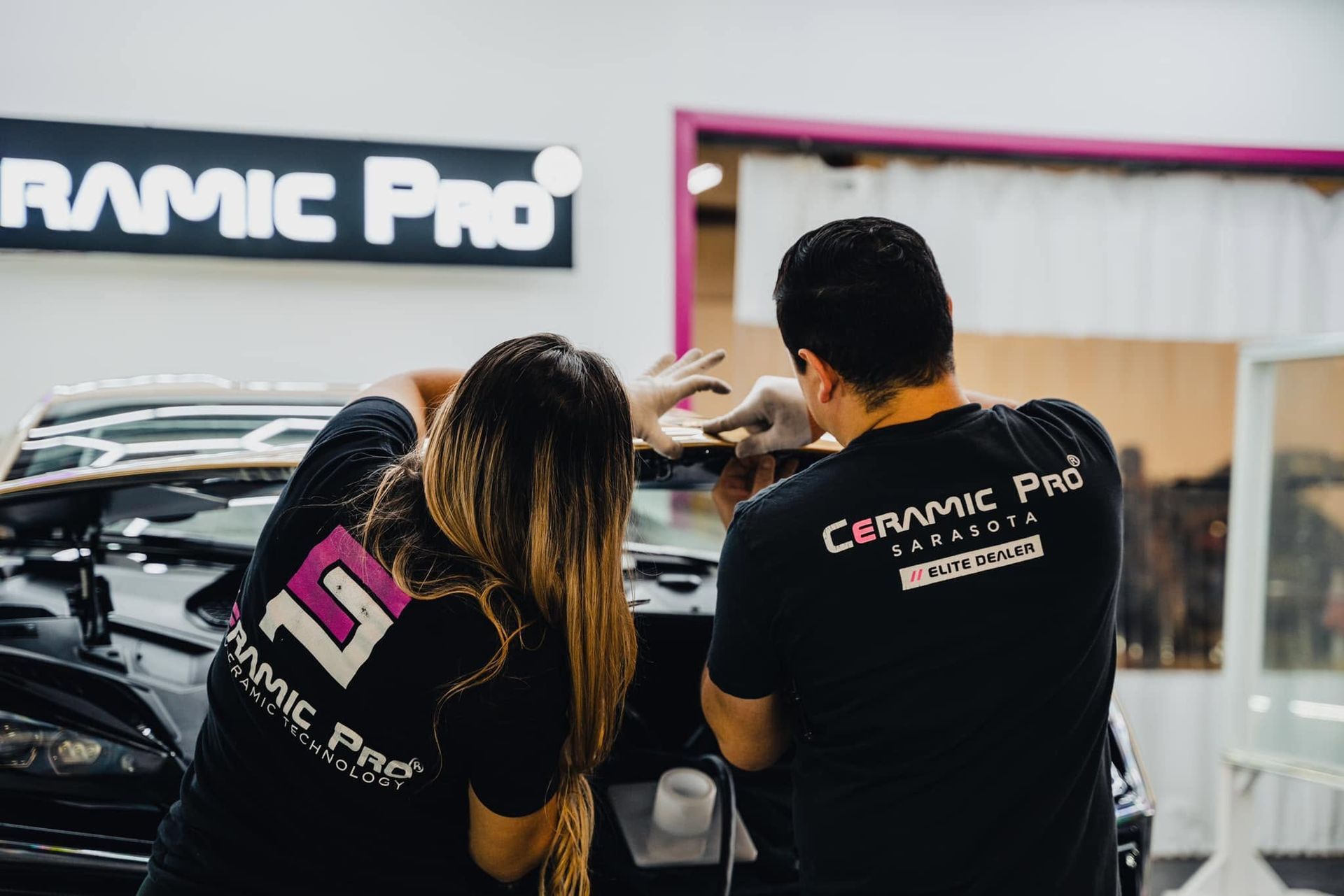 Two people in black Ceramic Pro shirts inspect a car in a showroom with the Ceramic Pro logo behind them.