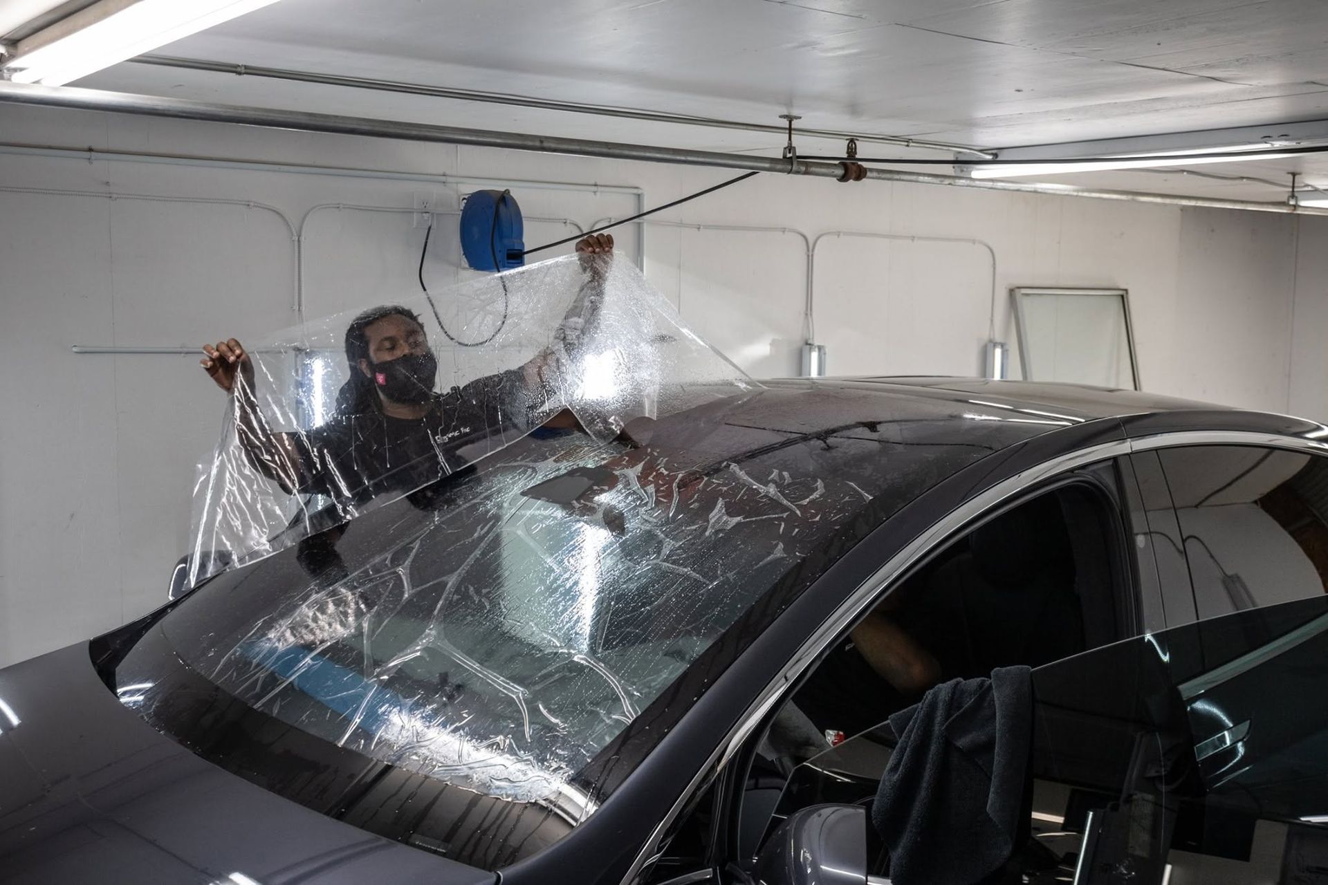 Person using plastic wrap over a black car in a garage during window or paint protection work