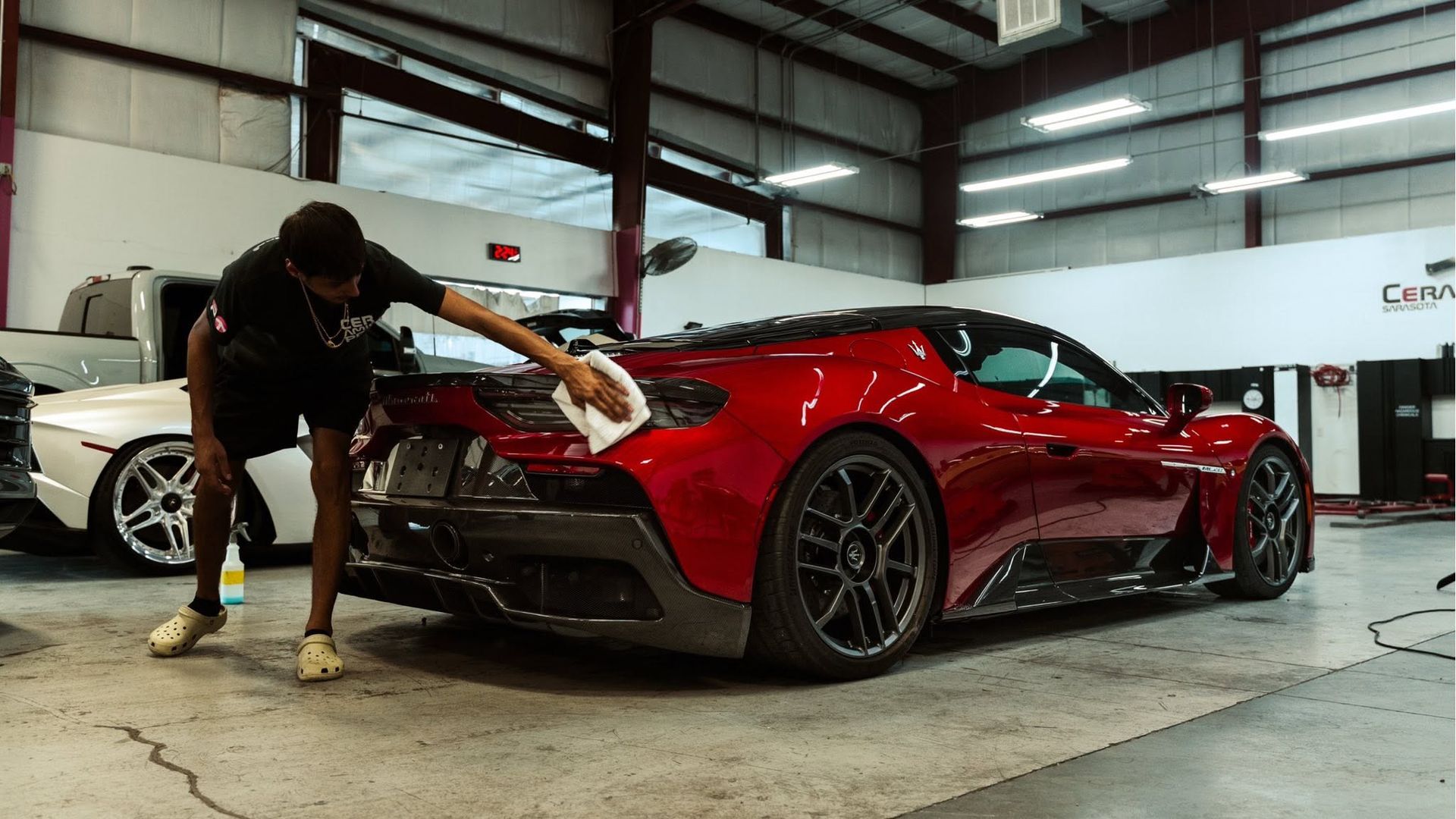 Person polishing a red sports car in a garage, with a white car in the background.