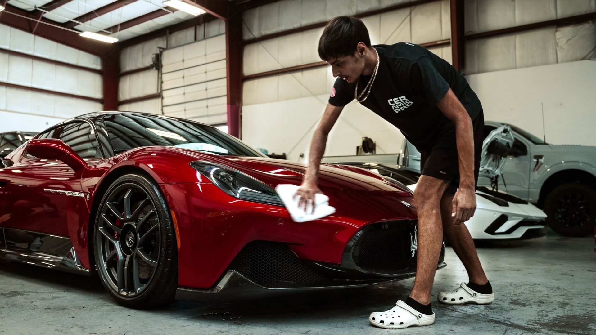 Man polishing a red sports car in a garage with a white car in the background