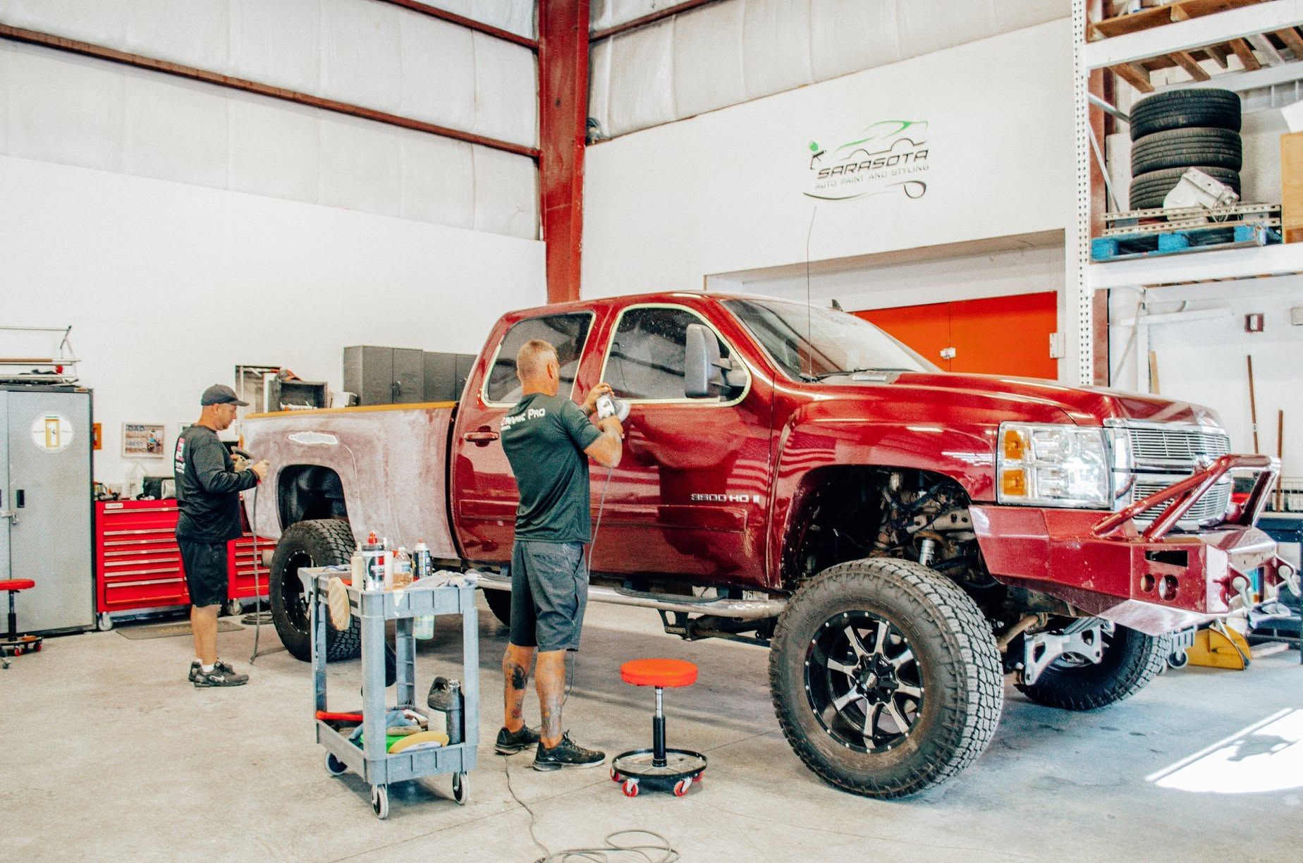 Red lifted pickup truck in a garage with two mechanics working on it.