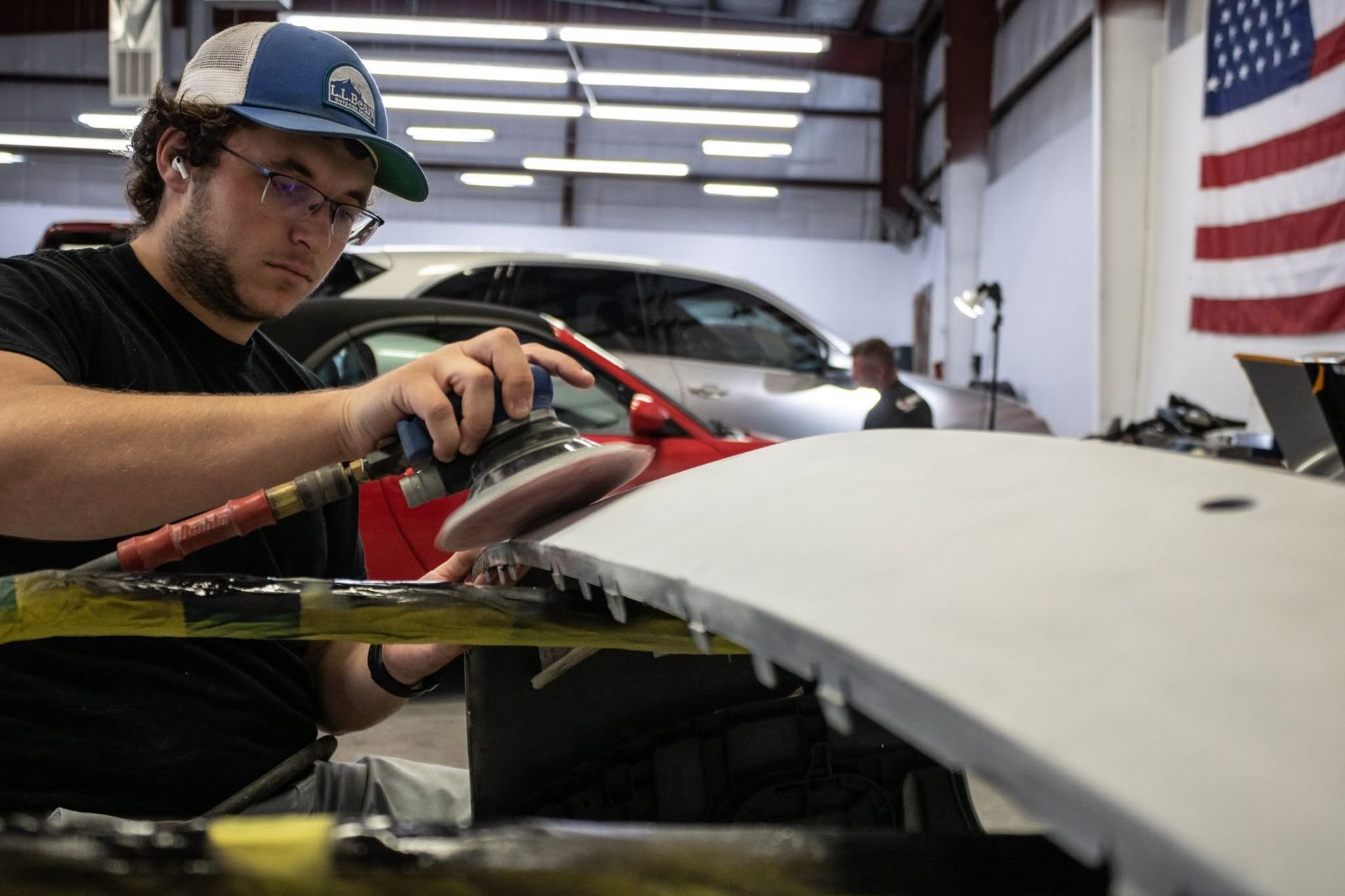 Worker sanding a white car panel in a workshop with a U.S. flag in the background