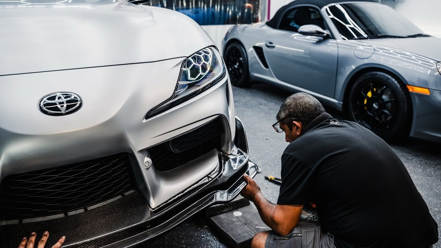 Mechanic working on the front bumper of a white Toyota sports car beside a silver coupe in a garage